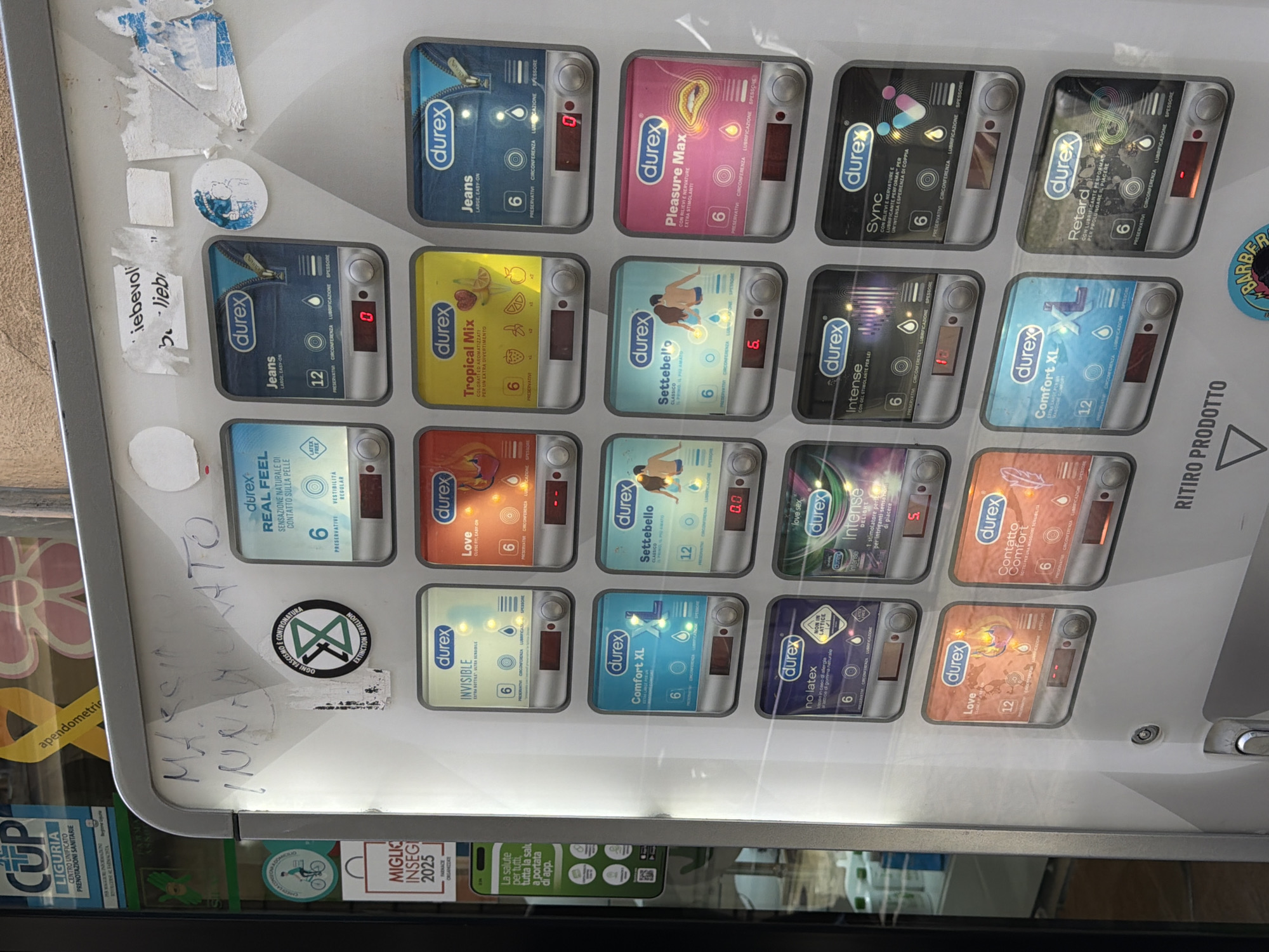The image shows a condom vending machine in what appears to be Genoa, Italy. The machine is stocked with various Durex condom types, including Jeans, Pleasure Max, Tropical Mix, Settebello, Intense, Comfort XL, Invisible, Love, and Sync. The vending machine's display shows that some selections are out of stock, indicated by a red "0" next to the product. Stickers and graffiti cover portions of the machine. A "MIGLIO INSEGI 2025" sticker and a "CUP LIGURIA" sticker are visible, as well as a sticker with a bicycle logo. The reflection in the machine's glass panel reveals a glimpse of the surrounding environment, including a glimpse of a shop window.