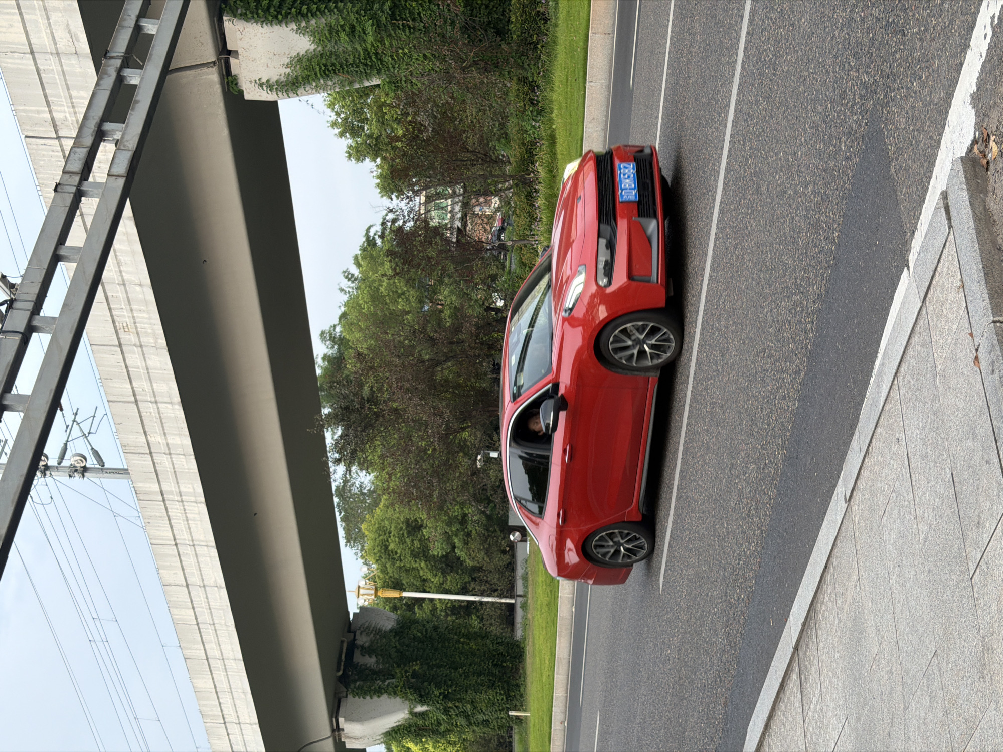 A red sedan is captured in motion on an asphalt road in Jiangjin, China. The car, with a license plate reading "BD BK582", has its front right wheel turned slightly inward, indicating a left turn or lane change. A person is visible in the driver's seat, looking forward. The road is bordered by a grey tiled sidewalk and a grassy area with trees and ivy-covered concrete structures. Overhead, a concrete overpass with exposed rebar and electrical wires is visible against a cloudy sky. The scene appears to be a typical urban environment with a focus on road infrastructure and a passing vehicle.