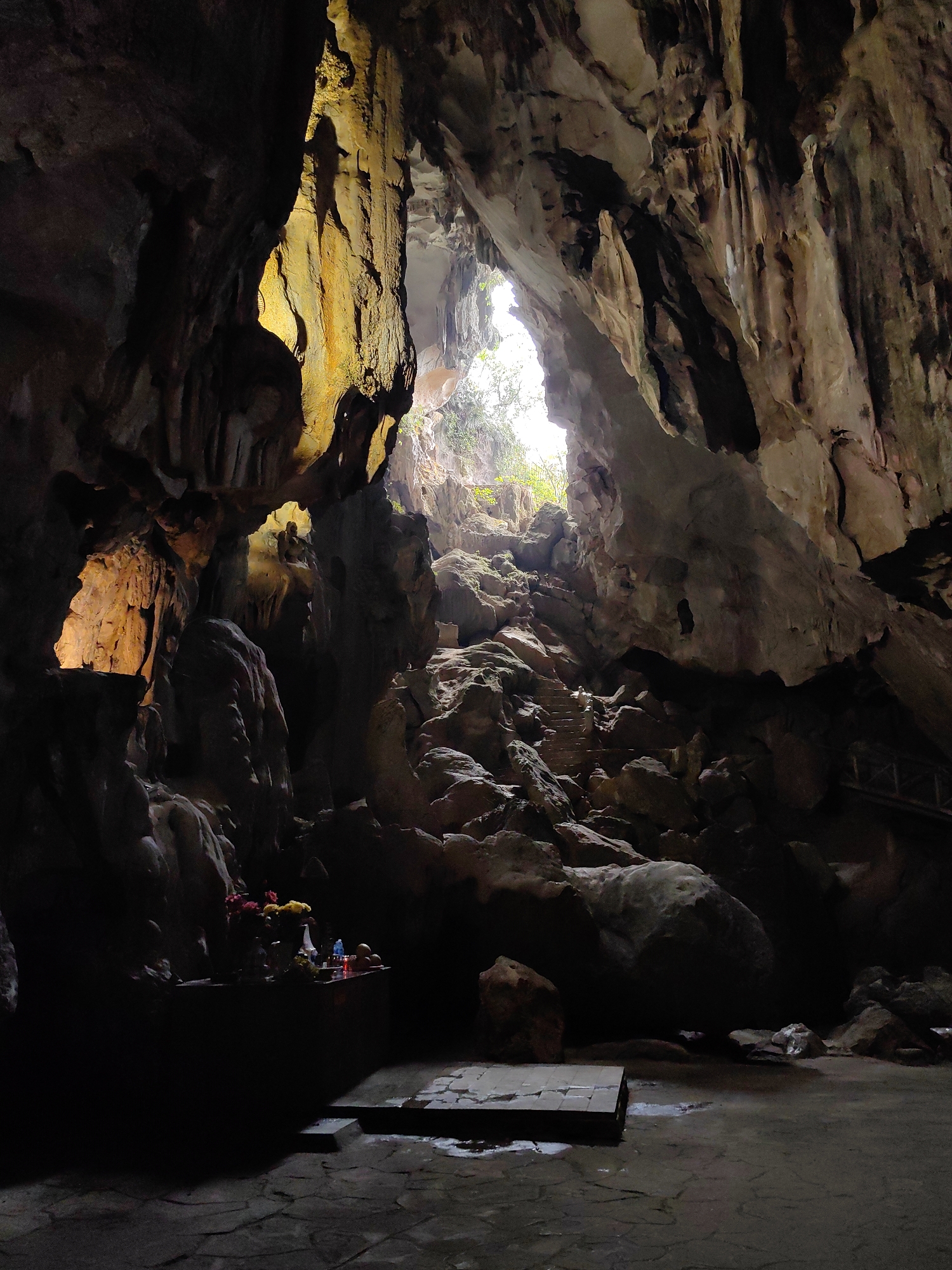 The image depicts the interior of a cave in Lạng Sơn, Vietnam. A large opening at the top allows daylight to filter in, illuminating a pile of rocks and stairs that lead towards the opening. The cave walls are rough and textured with formations. At the bottom left, a small altar with flowers, candles, and other offerings sits on a platform. The floor is paved with stone slabs.