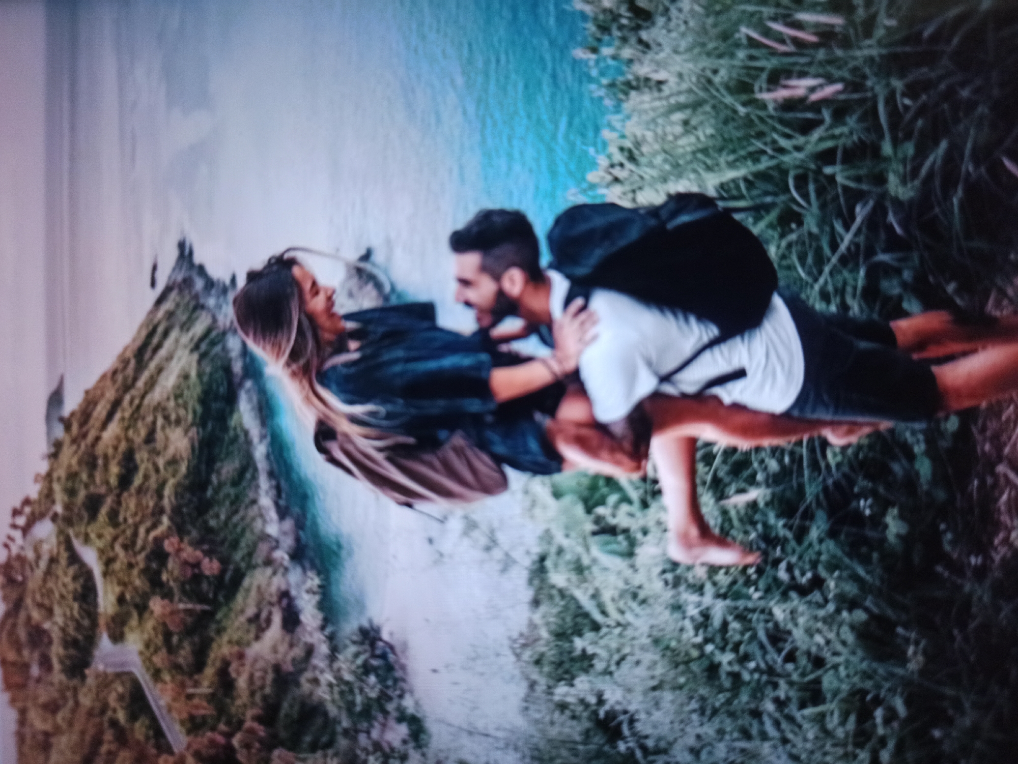 A man is holding a woman in his arms while standing on a grassy hill overlooking a body of water. The man is wearing a white t-shirt and dark shorts, and a black backpack. The woman is wearing a dark top and a light-colored backpack, and has her legs wrapped around the man. The background shows a coastline with lush green vegetation and a winding road. The water is a vibrant turquoise color, and the sky is a soft purple and blue gradient. The overall scene depicts a romantic and adventurous moment between a couple in a scenic natural setting. The location is not Balakliya, Ukraine; it is a coastal mountainous area with abundant greenery and ocean views.