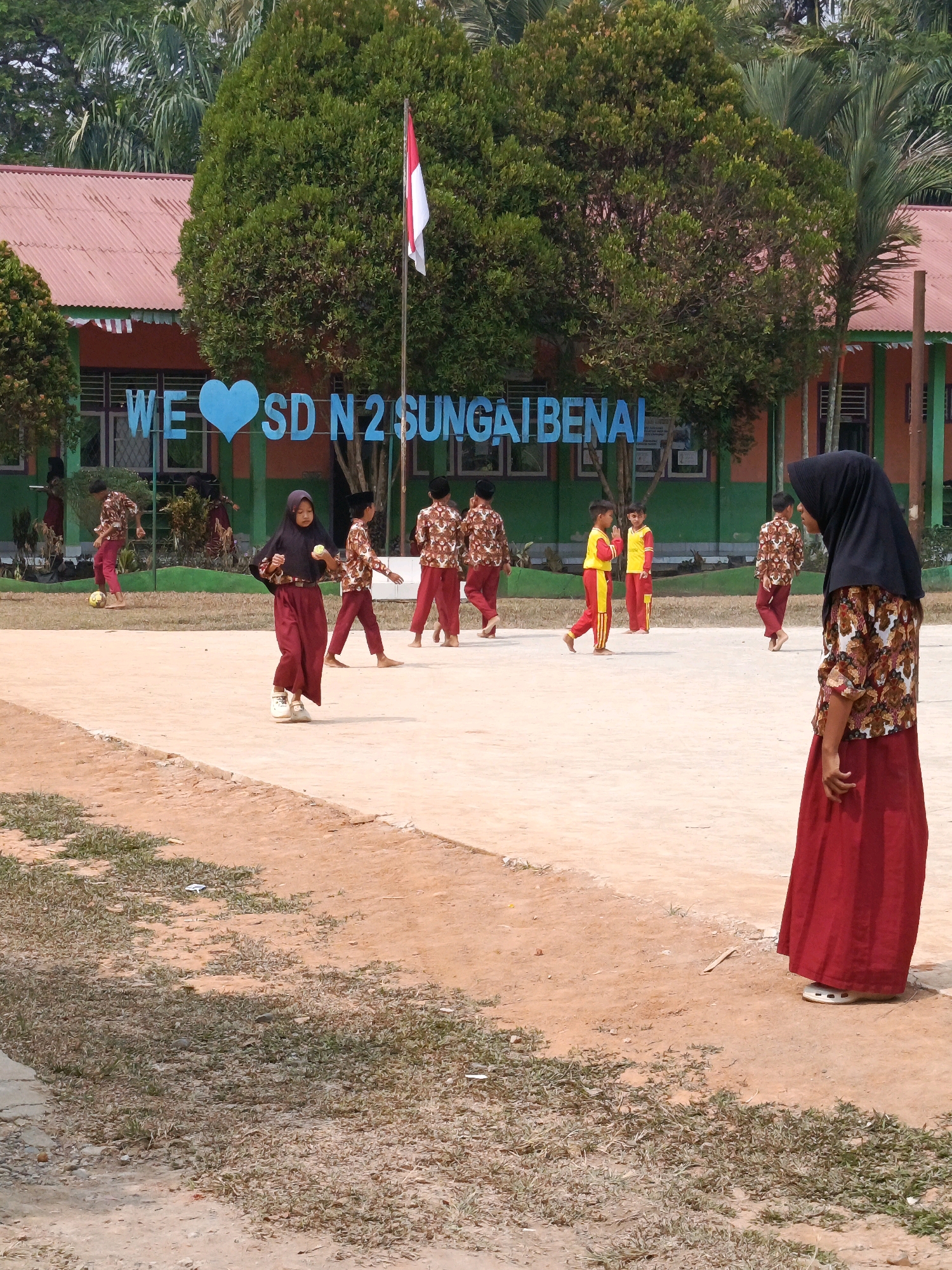 In Megang Sakti IV, Indonesia, a group of children are seen in the foreground on a sandy, outdoor court in front of a school building. A large, light blue sign with white lettering, reading "WE ♡ SD N 2 SUNGAI BENAI," is prominently displayed in front of the school. The school building has a red corrugated metal roof and orange walls with green trim. A flag with red and white stripes is flying on a pole near the building. Some children are wearing uniforms consisting of maroon pants and patterned shirts. One girl in the foreground is wearing a black headscarf and a maroon skirt and shirt, holding a yellow ball. Another girl in the far right is wearing a black headscarf, a patterned shirt, and a red skirt. In the background, palm trees and other lush vegetation surround the school. The scene appears to capture a school event or activity.