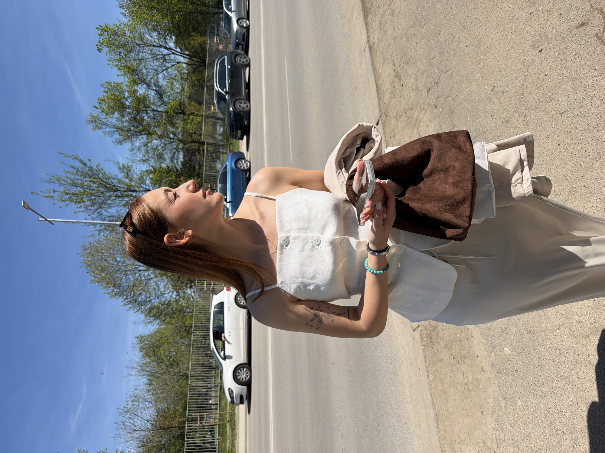 A young woman stands on a sidewalk in Sofia, Bulgaria, facing toward the sun. She is wearing a light-colored, sleeveless dress with a buttoned top. She holds a brown suede bag and a beige jacket. Her right arm has a tattoo. She is accessorized with a turquoise bracelet. Several cars are parked along the street, including a white van behind a metal fence. The sky is blue with green trees visible in the background. The location is in the city of Sofia, Bulgaria.