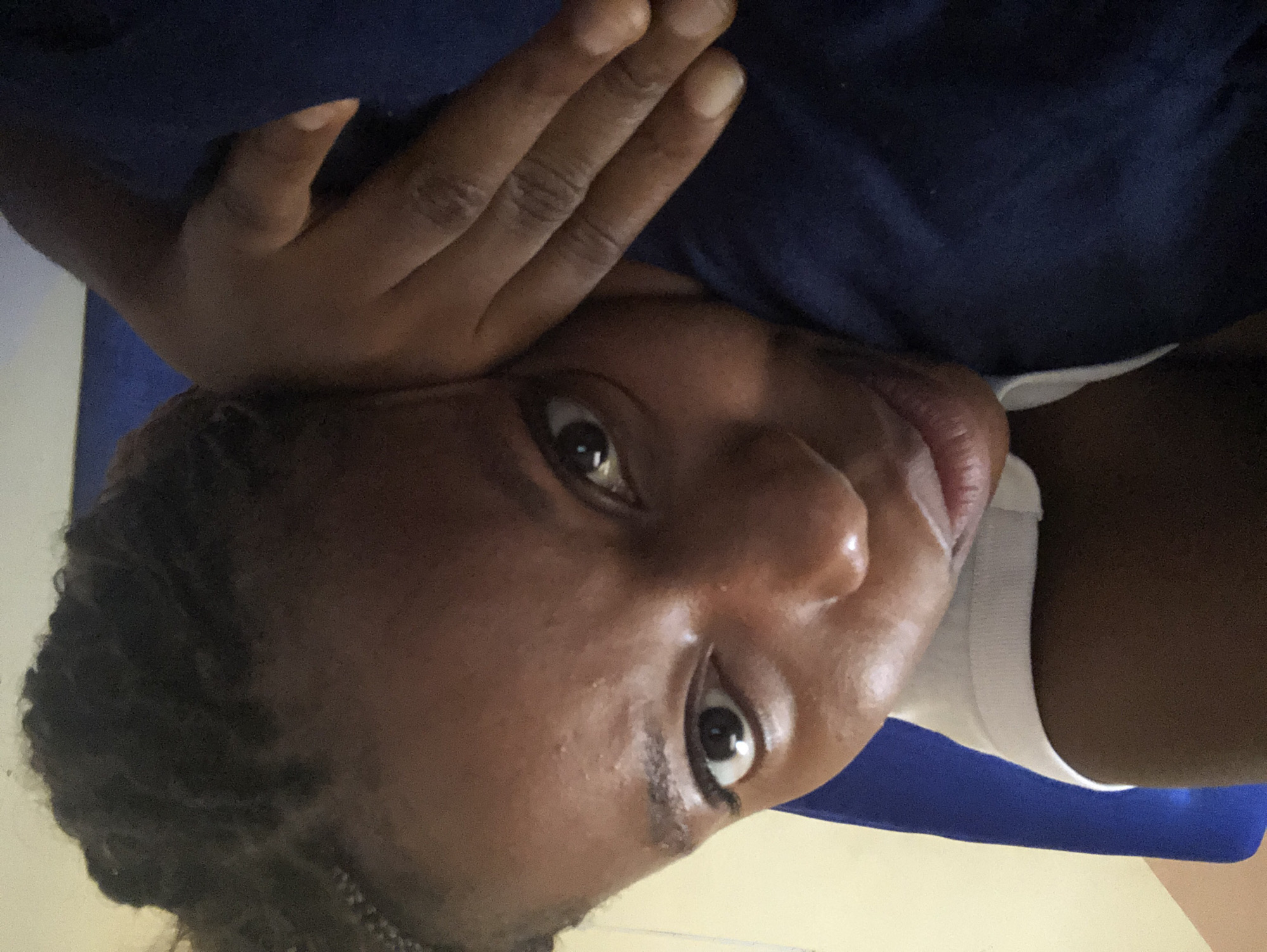 A close-up shot of a dark-skinned person with their hand partially obscuring their forehead. They have dark hair, and their eyes are open, looking directly at the viewer. The person is wearing a white garment. A blue cloth is visible in the background. Location is not discernible from the image.