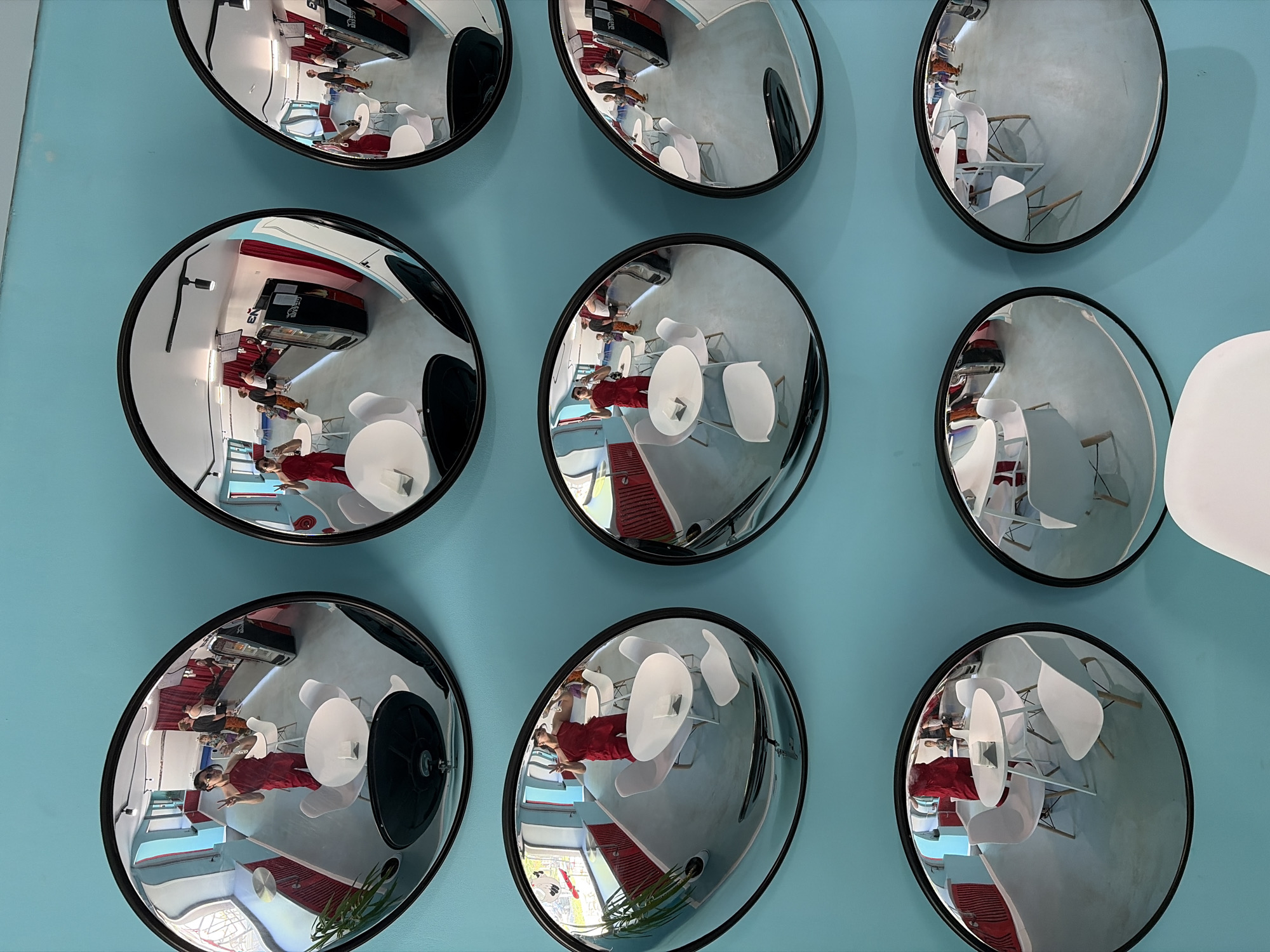 The image displays a blue wall decorated with nine round, black-framed convex mirrors arranged in a 3x3 grid. Each mirror reflects a partial view of a brightly lit interior, possibly a cafe or restaurant. Reflections include people seated at white tables with white chairs, a display case with a red accent, and other architectural details. A white object, possibly a piece of furniture, is partially visible in the lower right of the image, near the last mirror in the second row. The reflections are distorted due to the convex nature of the mirrors, and the orientation of the mirrors is varied, providing different perspectives of the interior. The location is Unknown, Unknown.