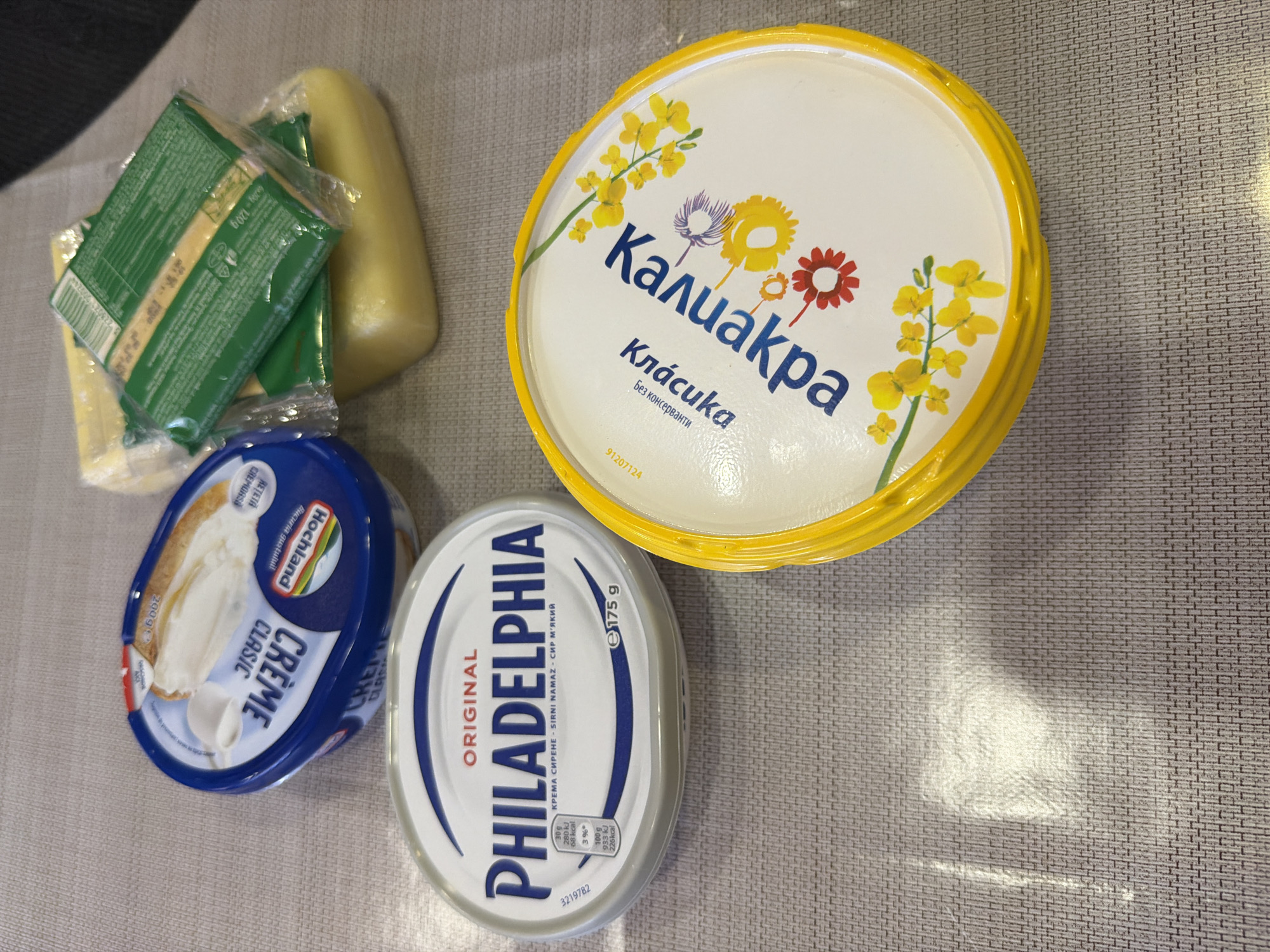 The image shows a top-down view of various dairy products arranged on a textured grey surface. In the foreground, a round, yellow-lidded container with a white label and yellow floral designs is prominently displayed. The label features blue Cyrillic text. To the left of this container is an oval, white and silver tub labeled "PHILADELPHIA ORIGINAL". To the left of that, a blue and white round container is visible, with the word "CREME" in large white letters on its lid. Behind the blue and white container, a block of yellow cheese wrapped in plastic and green foil packaging is partially visible. The overall setting appears to be an indoor environment, possibly a kitchen counter or table. There are no people visible in the image. The location of Novi Iskar, Bulgaria, is not visually identifiable from the provided image.