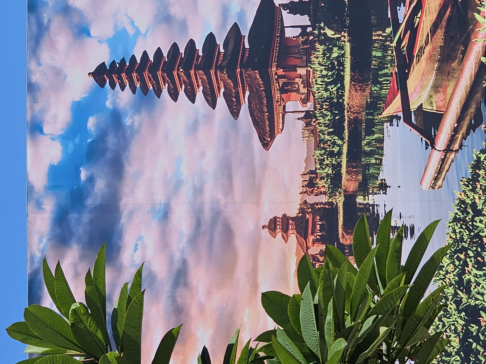 The image depicts a scenic view of the Pura Ulun Danu Beratan temple complex, located in Denpasar, Indonesia, situated on the shores of Lake Beratan. In the foreground, lush green foliage frames the left side of the scene, with large, broad leaves. The middle ground features a body of water reflecting the sky and the temple. A traditional Balinese boat, with its distinctive outriggers and colorful painted elements, rests on the water. Behind the water, the temple complex is visible. The main structure is a multi-tiered meru tower, characteristic of Balinese Hindu temples, with dark, layered roofs. To the left, a smaller, similar temple structure stands. The background consists of a dramatic sky with swirling clouds, illuminated by the warm hues of sunset or sunrise, casting a pink and orange glow across the scene. The overall impression is one of tranquility and cultural richness.