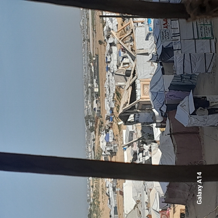 A high-angle shot reveals a sprawling refugee camp stretching into the horizon. Tents and temporary shelters cluster densely, framed by a dusty landscape. Beyond the camp, a distant settlement fades into the hazy sky. Visible through a window frame, the scene suggests observation from an elevated vantage point. The image conveys the scale and density of the refugee camp.