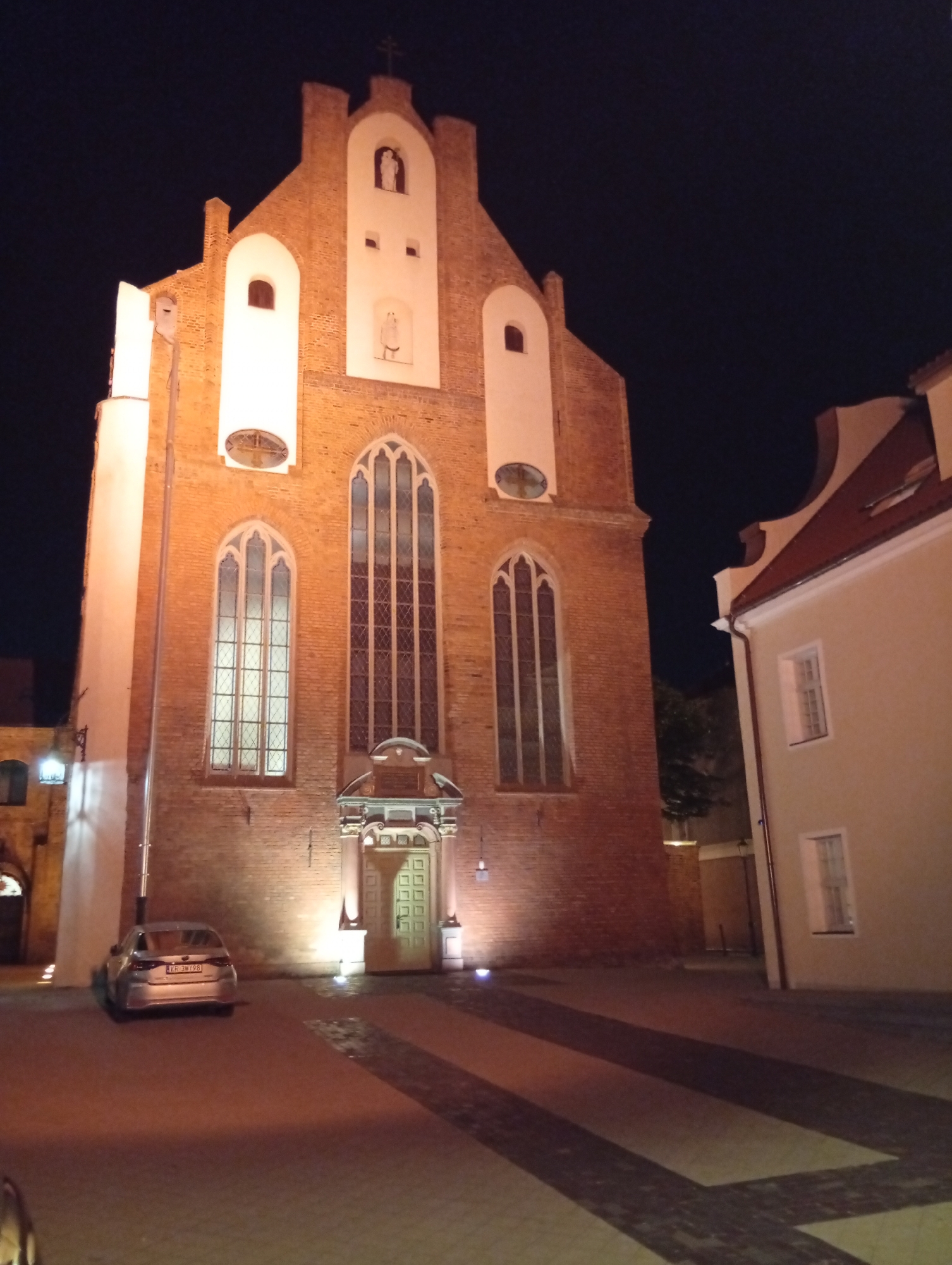 The image shows the exterior of St. Catherine's Church, a historic church in the city of Gdańsk, Poland, at night. The church is made of brick and features tall, Gothic-style windows and a prominent facade with statues. The scene is illuminated by artificial lighting, casting warm glows on the brickwork and surrounding paved area. A silver car is parked in front of the church, and a portion of a neighboring building with a red-tiled roof is visible to the right. The overall atmosphere is one of architectural grandeur and nocturnal serenity.