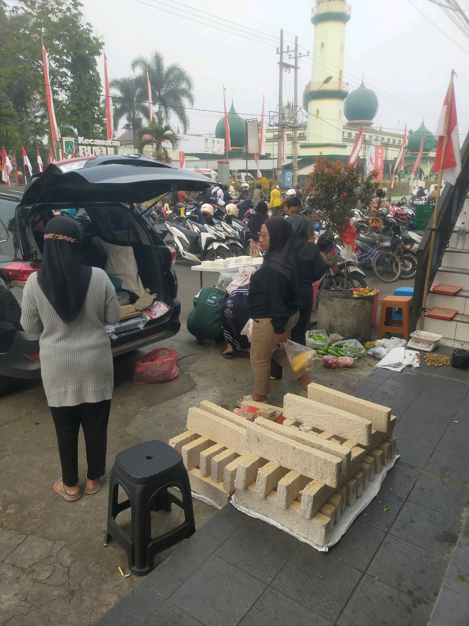 In Bululawang, Indonesia, a street scene unfolds with a mosque in the background, its green domes and tall minaret visible against an overcast sky. Numerous red and white Indonesian flags are displayed, some on tall poles and others attached to buildings. In the foreground, a black SUV with its trunk open is parked, with two women standing nearby. One woman, wearing a grey sweater and black hijab, stands facing the open trunk. The other woman, in a black hoodie and brown pants, is walking away from the car, carrying a plastic bag. A stack of rectangular, light-colored blocks, possibly food items like tempeh, is laid out on a white cloth on the pavement. Several motorcycles are parked in the mid-ground, and other individuals are visible in the background. The ground is paved with gray tiles. Scattered on the ground are various items including produce like cucumbers and onions, a red plastic bag, and a small black stool. A scale is also visible near the stack of blocks.
