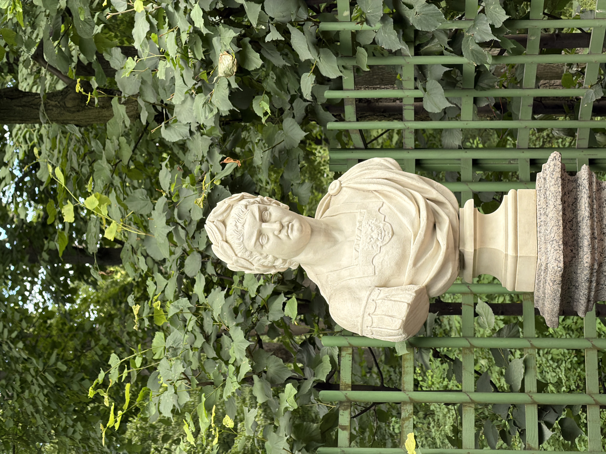 This is a full-shot, eye-level, outdoor photograph of a marble bust of a man wearing a laurel wreath and Roman armor. The bust is placed on a stone pedestal. Behind the bust, a green metal trellis is visible, with lush green foliage and trees forming a dense backdrop. The lighting suggests it is daytime, possibly overcast or with dappled sunlight filtering through the leaves. The overall scene appears to be in a park or garden. This sculpture is located in the Summer Garden in Saint Petersburg, Russia, specifically in the alley leading to the "Summer Palace".