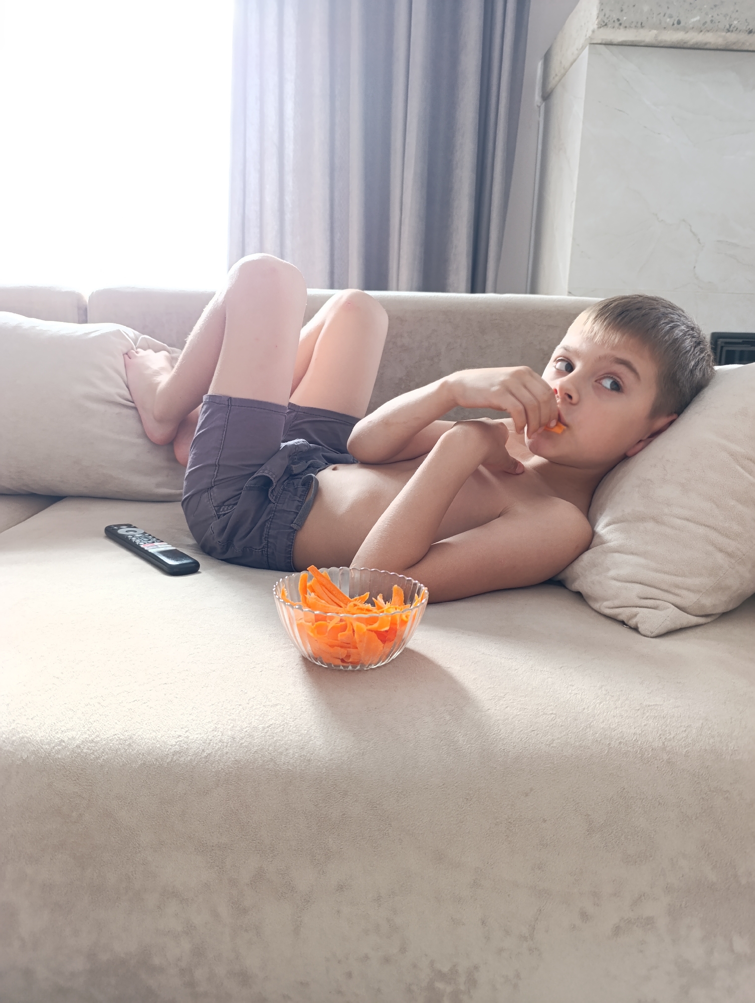 A shirtless boy lies on a light tan couch in a room with a window covered by sheer curtains. The couch has two pillows behind him and his bare feet rest on one of the pillows. He is wearing grey shorts. A bowl of orange carrot shavings is placed on the couch in front of him, and he is holding a shaving to his mouth. A black remote control rests on the couch near his waist. The boy is looking toward the camera. The location is Chaikovsky, Russia.