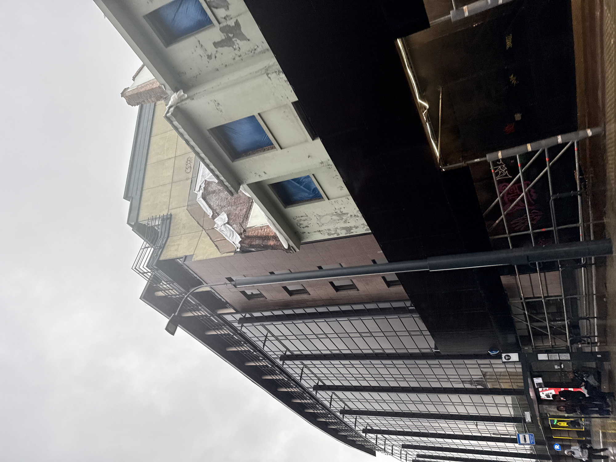 This image shows a street scene in Tallinn, Estonia. The foreground is dominated by a modern building with a glass facade, reflecting the overcast sky. To the right, an older building with peeling paint and blue tarps covering its windows is visible, appearing to be under renovation or demolition. Scaffolding is present in front of both structures, suggesting ongoing construction or repair work. A dark, covered walkway or entranceway with a glowing light is also visible in the lower right portion of the frame. A lamppost stands in front of the buildings. In the background, a few people are visible, some standing under an overhang, possibly waiting for transportation. A bus stop sign is mounted on the glass facade of the modern building. The ground appears wet, indicating recent rain. The overall atmosphere is urban and somewhat bleak due to the cloudy weather.