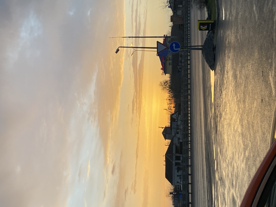 An eye-level shot captures a roadside view at sunset. The sky transitions from pale blue-grey to a radiant golden hue. Road signs and streetlights stand beside a wet road reflecting the sky. Buildings are silhouetted against the bright sky. A sliver of a vehicle's exterior appears at the frame's edge.