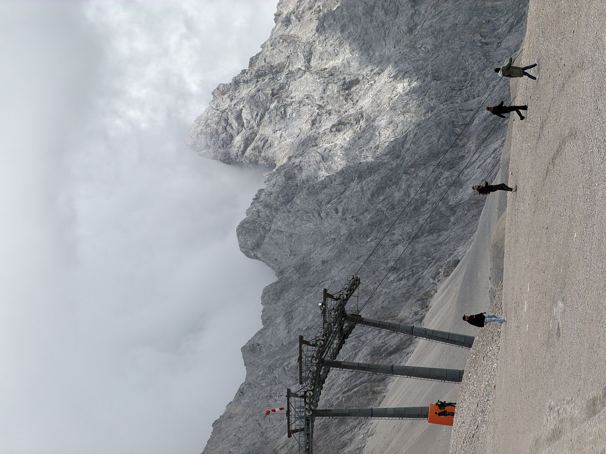 The image depicts a scenic mountain landscape with a ski lift structure in the foreground and rugged, rocky mountains in the background partially obscured by clouds. Several individuals are visible walking on a gravel path in front of the mountains. Two individuals stand near an orange structure and ski lift supports on the left side of the frame. Further to the right, three individuals are walking across the path. The sky is overcast with scattered clouds. This scene is likely from the Zugspitze mountain, the highest peak in Germany, located near Garmisch-Partenkirchen.