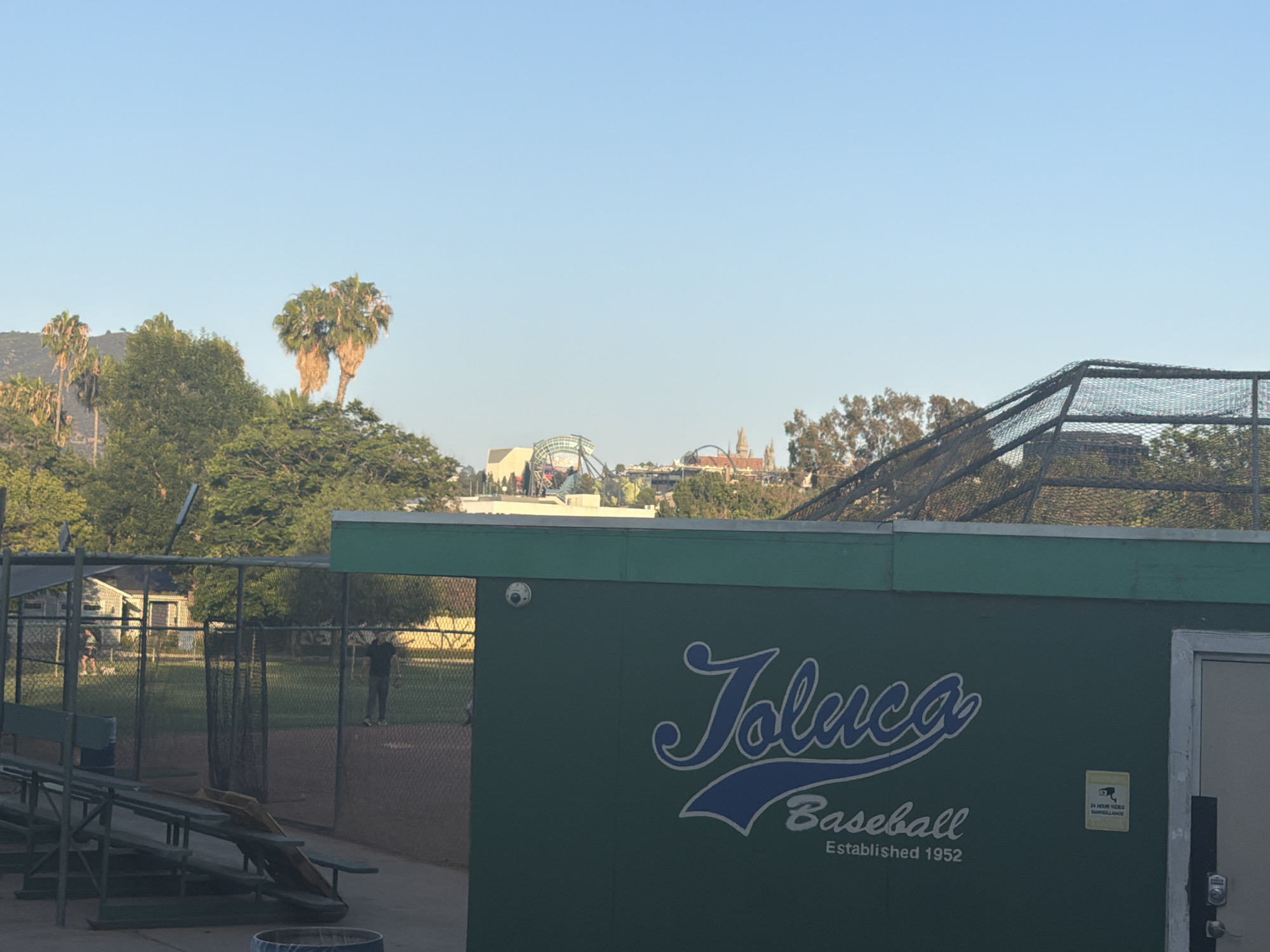 The image captures a view of a baseball field and its adjacent facilities, likely in a park-like setting within Los Angeles, USA. In the foreground, a green dugout structure is visible with the text "Toluca Baseball Established 1952" prominently displayed in white and blue lettering. A security camera is mounted on the wall of the dugout. To the left of the dugout, bleachers and a chain-link fence surround the baseball field, with a backstop visible behind home plate. In the background, several palm trees and dense green foliage are present, suggesting a warm climate. Beyond the trees, a cityscape with structures, possibly amusement park rides or buildings with distinctive architecture, can be seen under a clear blue sky. The overall scene suggests a well-maintained community sports facility.
