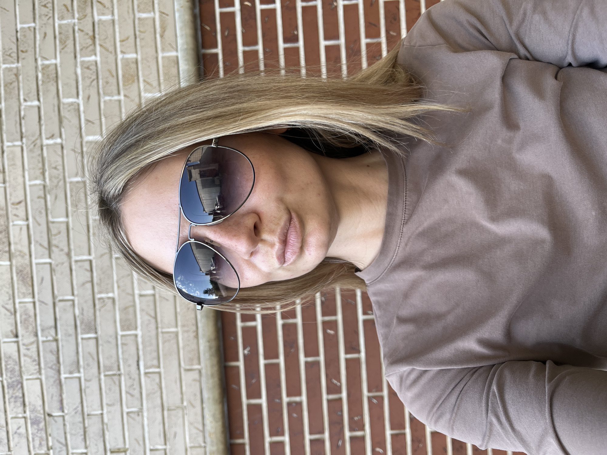 A close-up portrait shot depicts a person, likely female, from the shoulders up, with their head tilted. The person has straight, blonde or light brown hair and is wearing aviator-style sunglasses with reflective lenses. Their mouth is closed with a neutral to slightly pensive expression. They are dressed in a plain, long-sleeved shirt of an earth-toned color, possibly brown or taupe.
The background features a brick wall, distinctly divided into two sections: lighter, cream or beige-colored bricks on the left side and darker, reddish-brown bricks on the right side, suggesting a building corner or two adjacent walls. The bright lighting indicates it is daytime. Reflections in the sunglasses show parts of a building structure, suggesting an outdoor environment, possibly suburban or urban, in Chaikovsky, Russia. No specific activity is captured beyond the person posing. There is no visible text in the image.