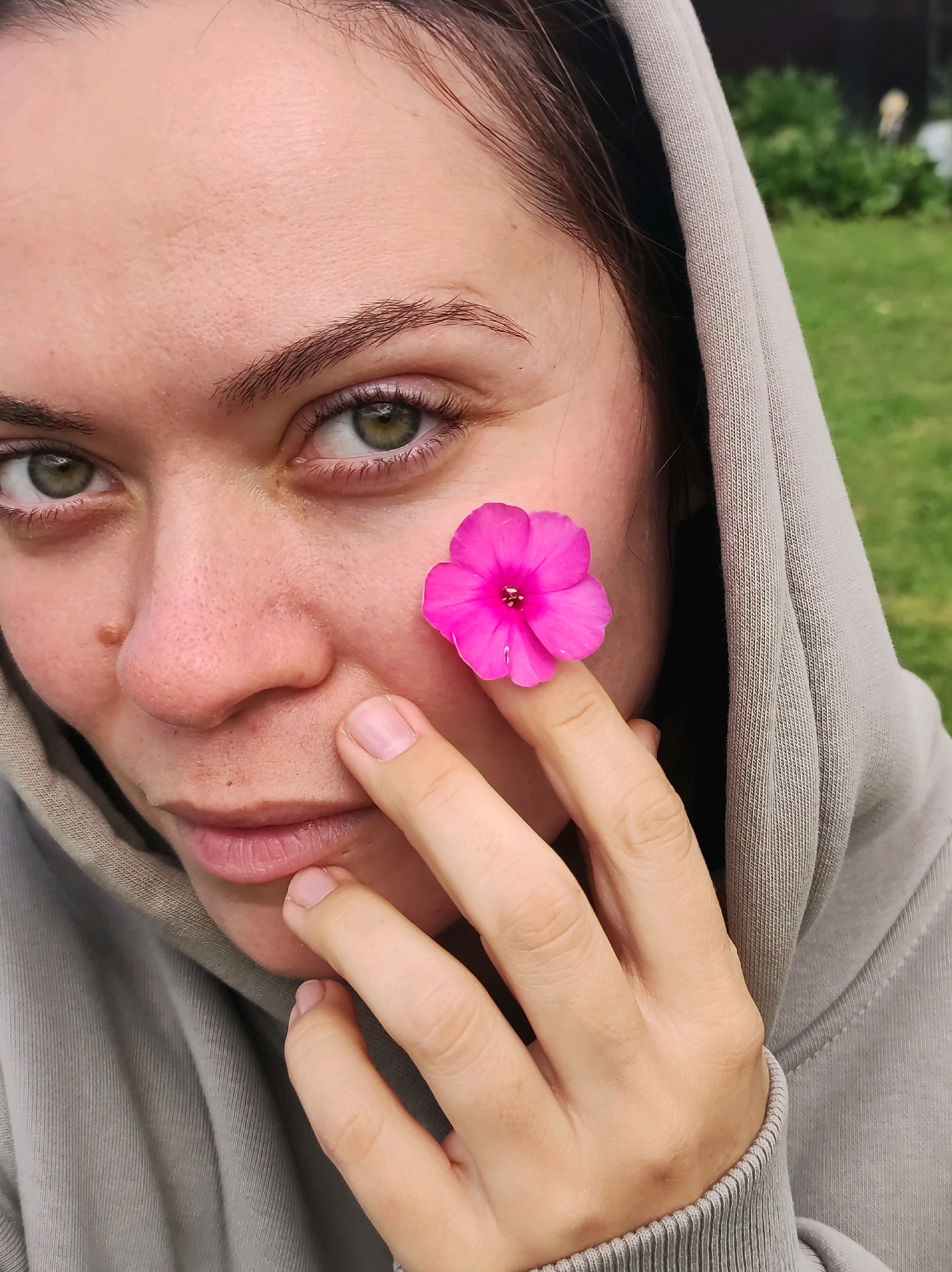 The image shows a close-up of a young woman's face, partially obscured by the hood of a grey sweatshirt. She has fair skin, green eyes, and dark hair. A small, bright pink flower is held against her cheek by her right index finger. Her left hand is near her chin. The background appears to be an outdoor setting with blurred green grass and foliage, suggesting a natural or park-like environment. The woman's gaze is directed towards the camera. There is no identifiable information to suggest the location is in Фоки, Russia.