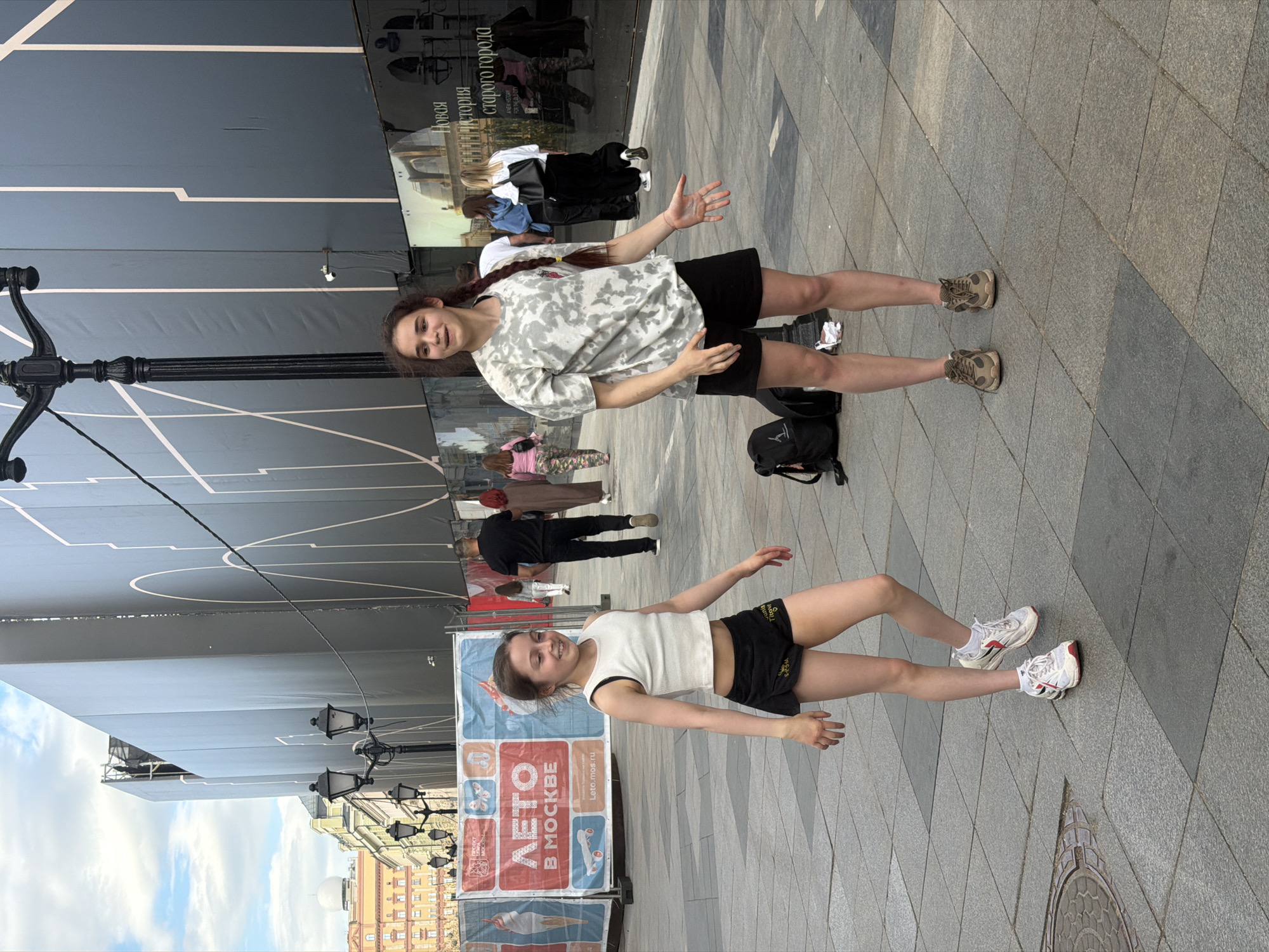 The image shows two young women standing on a public plaza in Moscow, Russia. Both women are wearing athletic clothing, shorts, and athletic shoes. One woman is wearing a white and gray t-shirt, while the other is wearing a white tank top. In the background are buildings, street lights, and a large poster board that reads, "ЛЕΤΟ В МОСКВЕ" ("Summer in Moscow"). The poster also includes the website leto.mos.ru. A sign or banner containing the text "Новая история старого города" (A new history for the old city) is also partially visible behind the women. The scene appears to be taken during the daytime. There are other people walking around in the background.