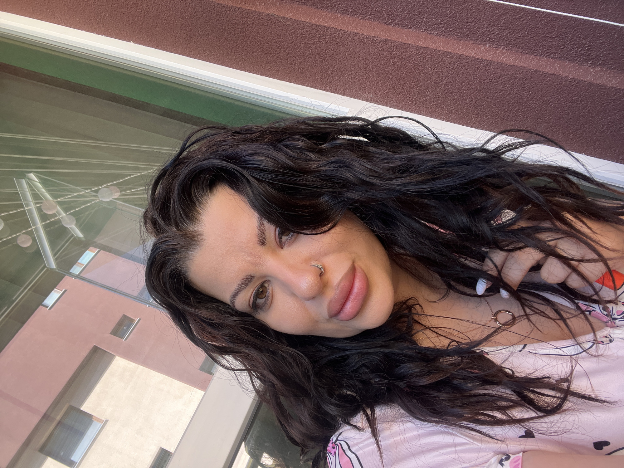 A woman with dark, wavy hair, light brown eyes, and a nose ring is facing the camera. She is wearing a pink patterned top. The background shows a window with a building reflection and a dark brown textured wall. Based on the architectural style visible in the reflection, this photo may have been taken in Plovdiv, Bulgaria, although it cannot be definitively determined without additional information.