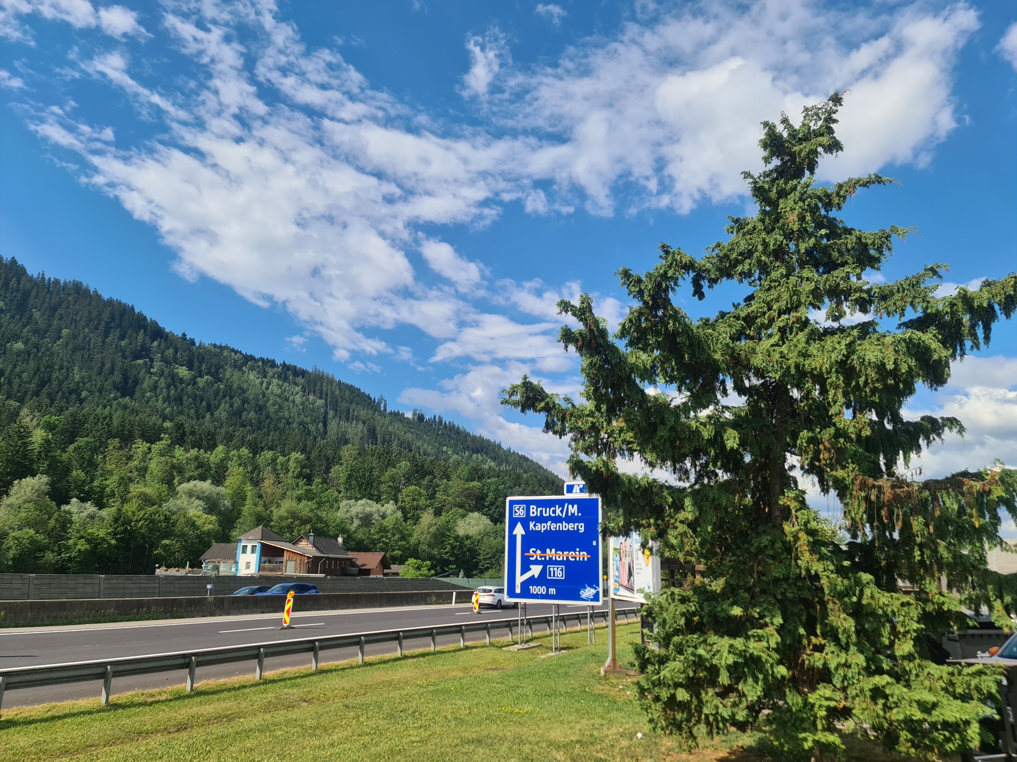 An eye-level outdoor medium shot captures a highway on a sunny day with a dense forest covering a mountainside. In the foreground to the right, a large evergreen tree partially obscures a blue directional sign. The sign indicates that "S6 Bruck/M. Kapfenberg" is straight ahead with an arrow pointing up, and "St. Marein 116" is to the right, with an arrow pointing right and the text "1000 m" below it. A white car is driving on the highway, and two dark blue cars are parked in front of buildings on the left side of the road. The buildings appear to be houses or small businesses. The highway has a concrete barrier on the left and a metal guardrail on the right. The sky is bright blue with scattered white clouds. The location is Sankt Marein im Mürztal, Austria.