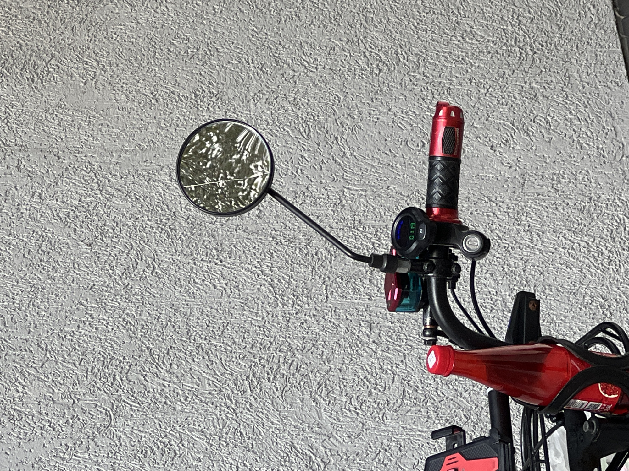 The image shows a close-up of the handlebars of a red electric scooter parked against a textured gray wall. A round mirror is attached to the left handlebar, reflecting trees and foliage. Below the mirror, a digital display reads "01:19". To the right of the display is a red and black grip. A red bottle, possibly of juice or soda, is secured in a black holder on the left side of the handlebars. The background wall appears to be an exterior surface of a building. There are no people or animals visible in the frame. The scene is an outdoor, urban environment, but no specific location within Moscow, Russia, can be determined from the visual information provided.