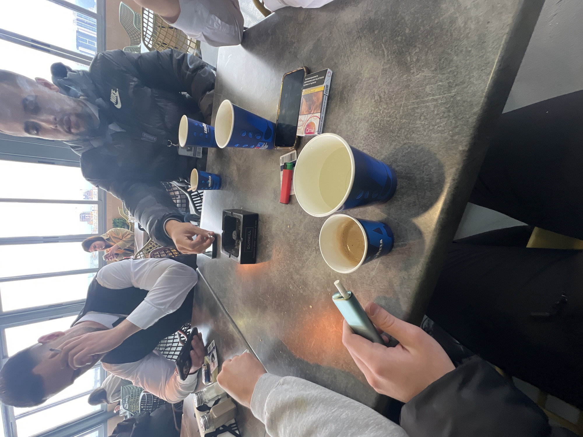 Here's a description of the image:
The image shows a group of people seated around a table in what appears to be an indoor cafe or restaurant setting. The location is likely in Sofia, Bulgaria, based on the Lavazza coffee cups on the table.
People:
* Several people are visible partially in the image. Two men are smoking. One of them is wearing a white dress shirt with a black vest, the other is wearing a black Nike down jacket.
* At least two other individuals are partially visible; one wearing a beige jacket.
* Hands are visible holding a blue electronic cigarette device.
Objects:
* Several blue Lavazza coffee cups are on the table, some full, some empty.
* A pack of Ome cigarettes and a lighter are present.
* A black smartphone.
* A black Dunhill ashtray.
* The table is made of gray, textured material.
Actions/Interactions:
* People are engaged in smoking cigarettes and the electronic cigarette.
* The overall scene suggests a casual social gathering.
Environment:
* The setting is indoors, likely a cafe or restaurant.
* Large windows in the background provide a view of a cityscape, suggesting a building with a high vantage point.
* The room has modern furniture and a casual atmosphere.