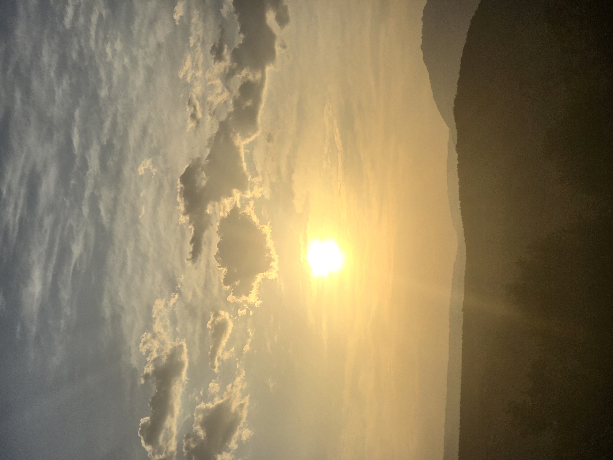 The image depicts a panoramic view of the outskirts of Podgorica, Montenegro, at sunset. The sky is filled with a mix of soft, wispy clouds in shades of grey and white, illuminated by the bright, golden-yellow disc of the sun. Rays of sunlight emanate from the sun, casting a warm glow across the scene and creating visible light shafts.
In the foreground, a silhouetted landscape of rolling hills and trees is visible, appearing dark and lush. The hills recede into the distance, with layers of mountainous terrain fading into the hazy twilight. The overall atmosphere is serene and natural, suggesting a rural or semi-rural setting just outside the city limits. There are no visible people or man-made structures, focusing solely on the natural environment and the dramatic interplay of light and shadow.