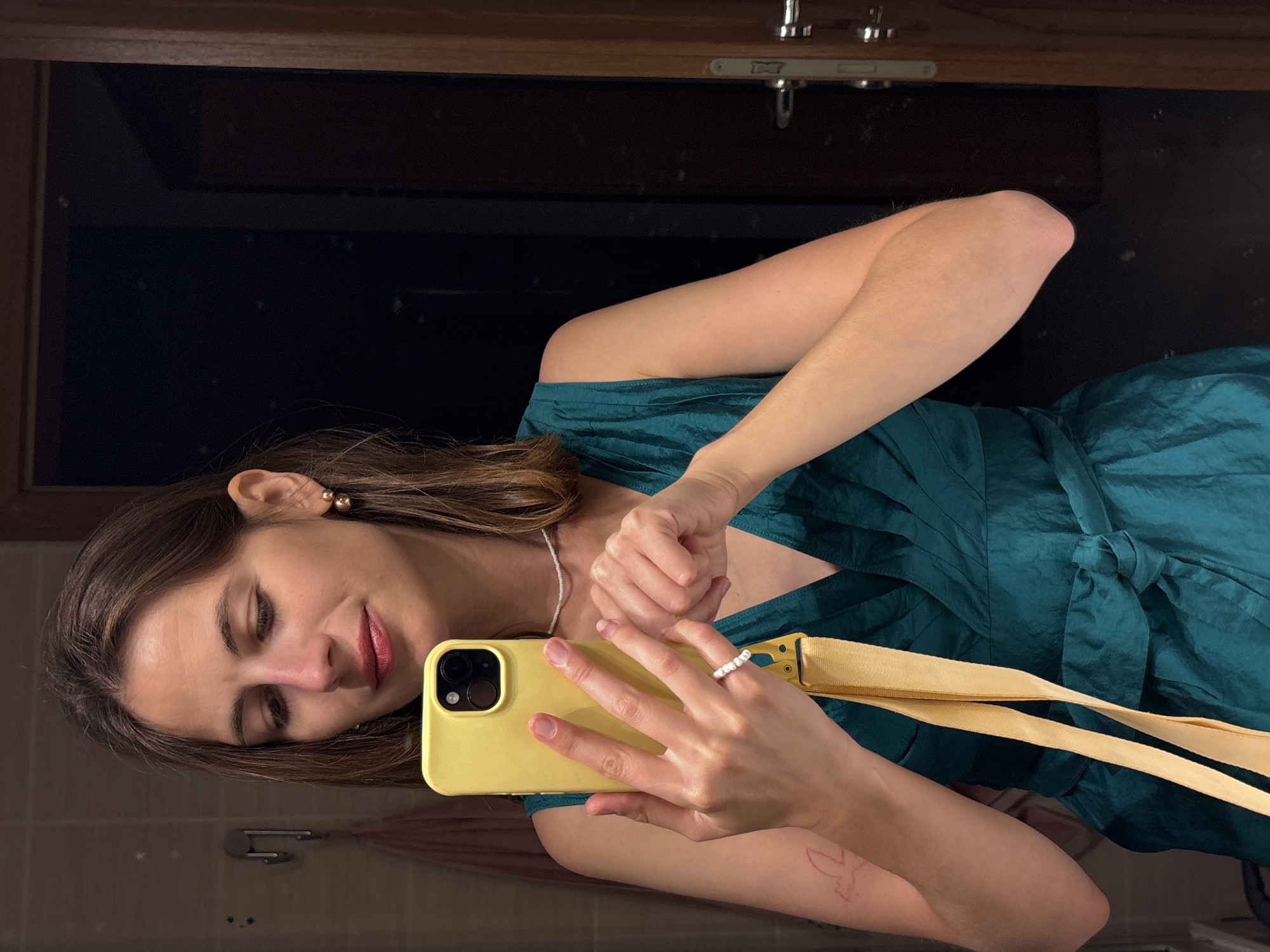 A young woman with brown hair is taking a selfie in a mirror. She is wearing a teal wrap dress and a pearl necklace. She has a yellow phone in her left hand and is wearing a white beaded ring on her right index finger. A red tattoo of a bird is visible on her left upper arm. The background appears to be a bathroom with a light-colored tiled wall and a wooden doorframe with a silver handle. The woman is positioned in front of a mirror, reflecting her image. The scene is indoors, likely in a private residence or hotel room in Bečići, Montenegro.