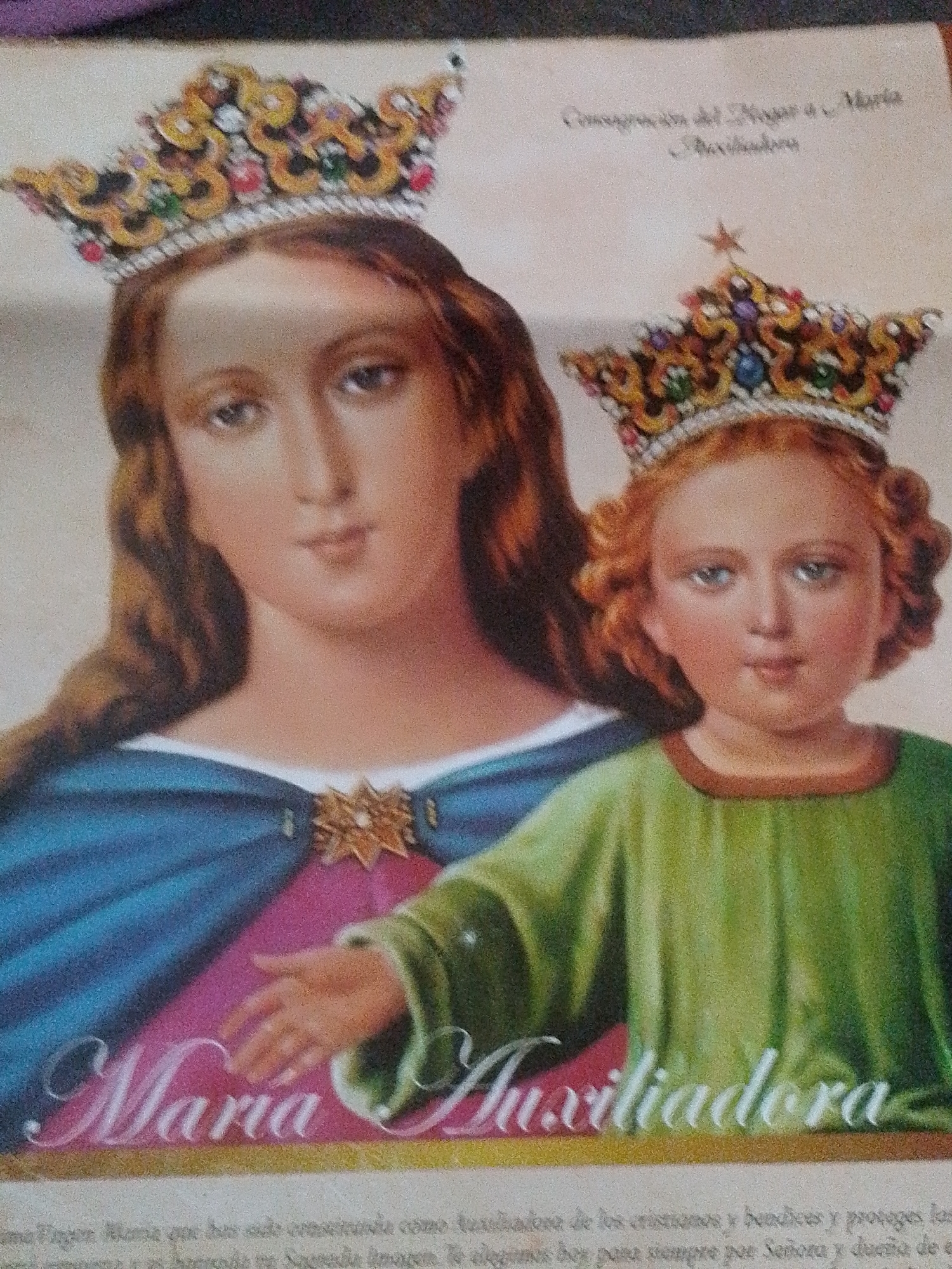 The image is a close-up photograph of a religious painting, likely an icon or a devotional image. It depicts the Virgin Mary and the Child Jesus, both wearing ornate crowns. The Virgin Mary is on the left, with her head tilted slightly and her eyes looking forward. She is wearing a blue mantle over a pink garment, fastened with a decorative brooch. The Child Jesus is on the right, also crowned, wearing a green tunic. He is reaching out his hand towards the viewer or towards his mother. Below the figures, the words "Maria Auxiliadora" are written in large, ornate script. The background is a light, textured color. There is also some faint text in the upper right and bottom of the image, which appears to be in Spanish. There is no discernible information within the image to suggest a specific location in Santa Ana, Venezuela.