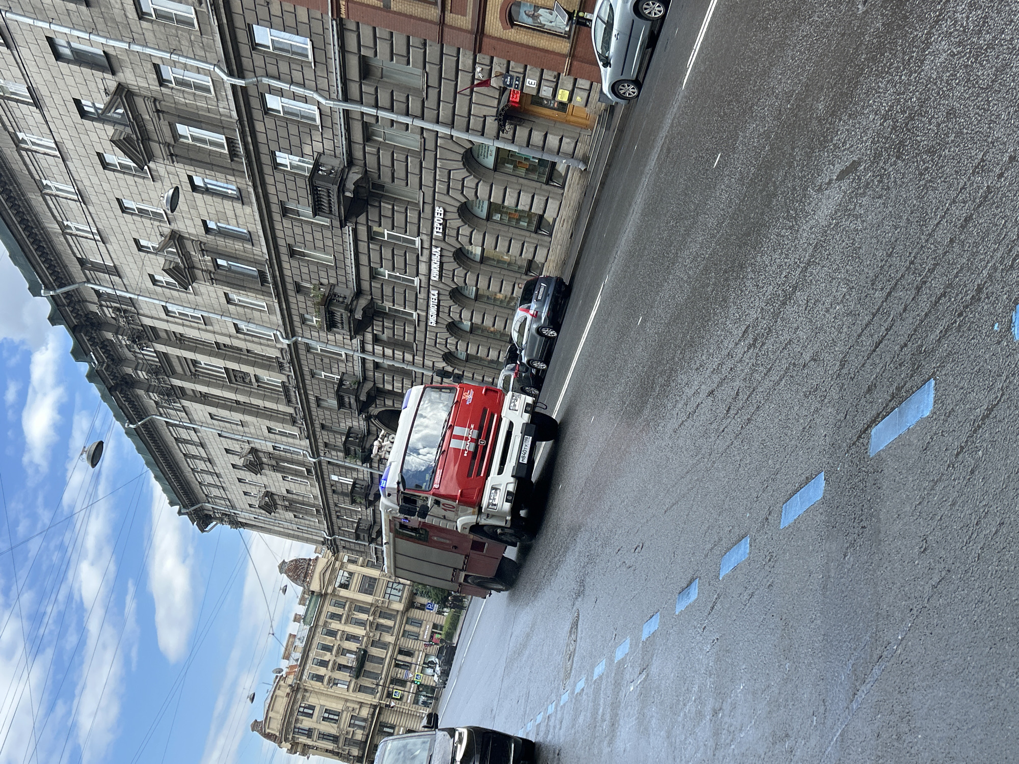 A red and white fire truck with the number 10 on its side is traveling down a wet city street. The fire truck has blue flashing lights on top. In the background, there are large, ornate, old buildings with many windows. There are also parked cars and some street lights. The sky is cloudy. The street has blue rectangular markings on it.