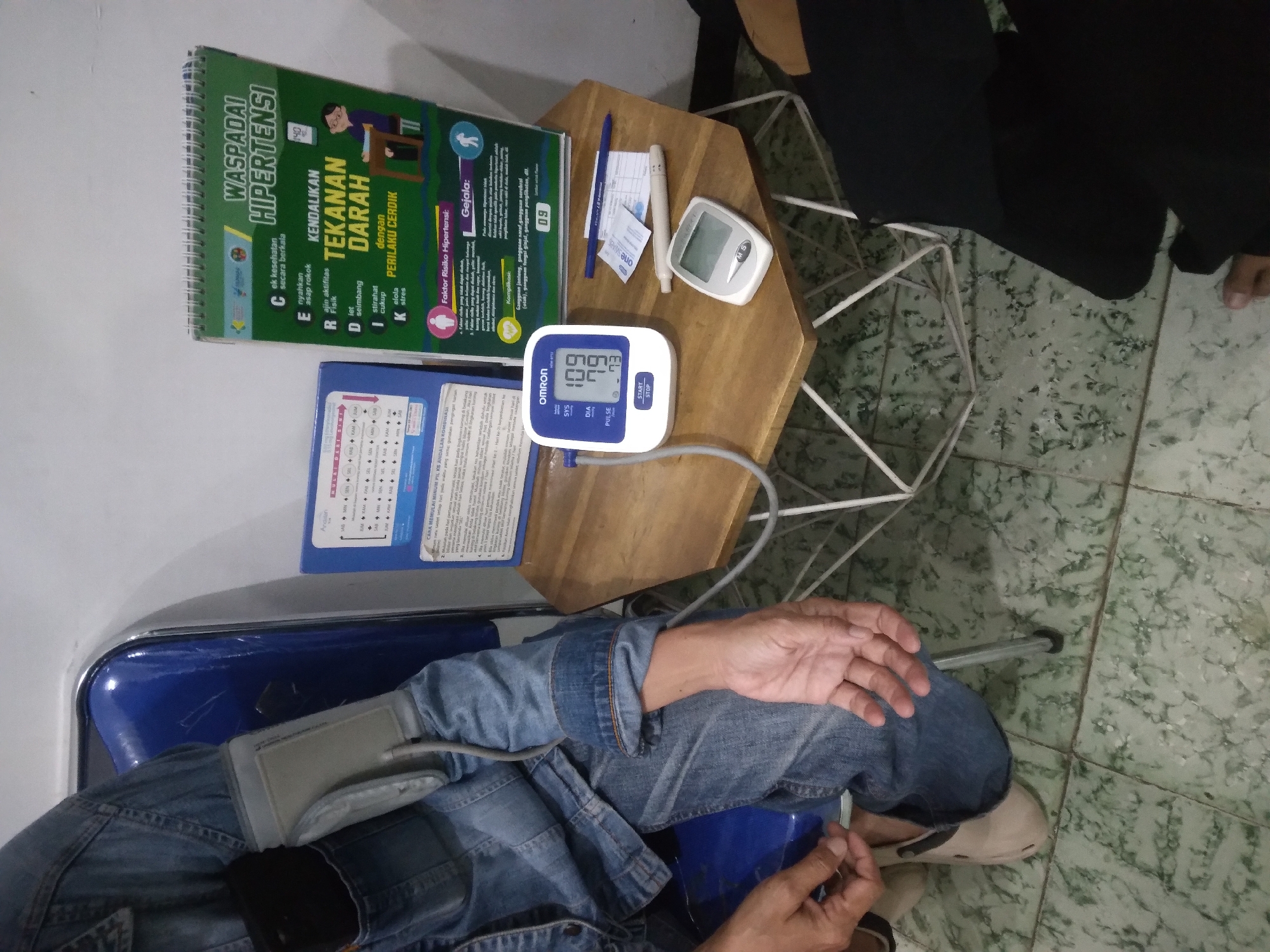 An individual is seated on a blue plastic chair in what appears to be a medical setting, possibly a clinic in Mersi, Indonesia. The person is wearing a denim jacket and jeans. Their left arm is extended, and a blood pressure cuff from an Omron blood pressure monitor is attached to it. The monitor displays readings of SYS 109, DIA 79, and PULSE 73. A glucometer and a pen are placed on a hexagonal wooden table next to the chair. In the background, a flip chart with the title "WASPADAI HIPERTENSI" (Beware of Hypertension) is visible, along with informational pamphlets. The floor is tiled with a green, marble-like pattern.