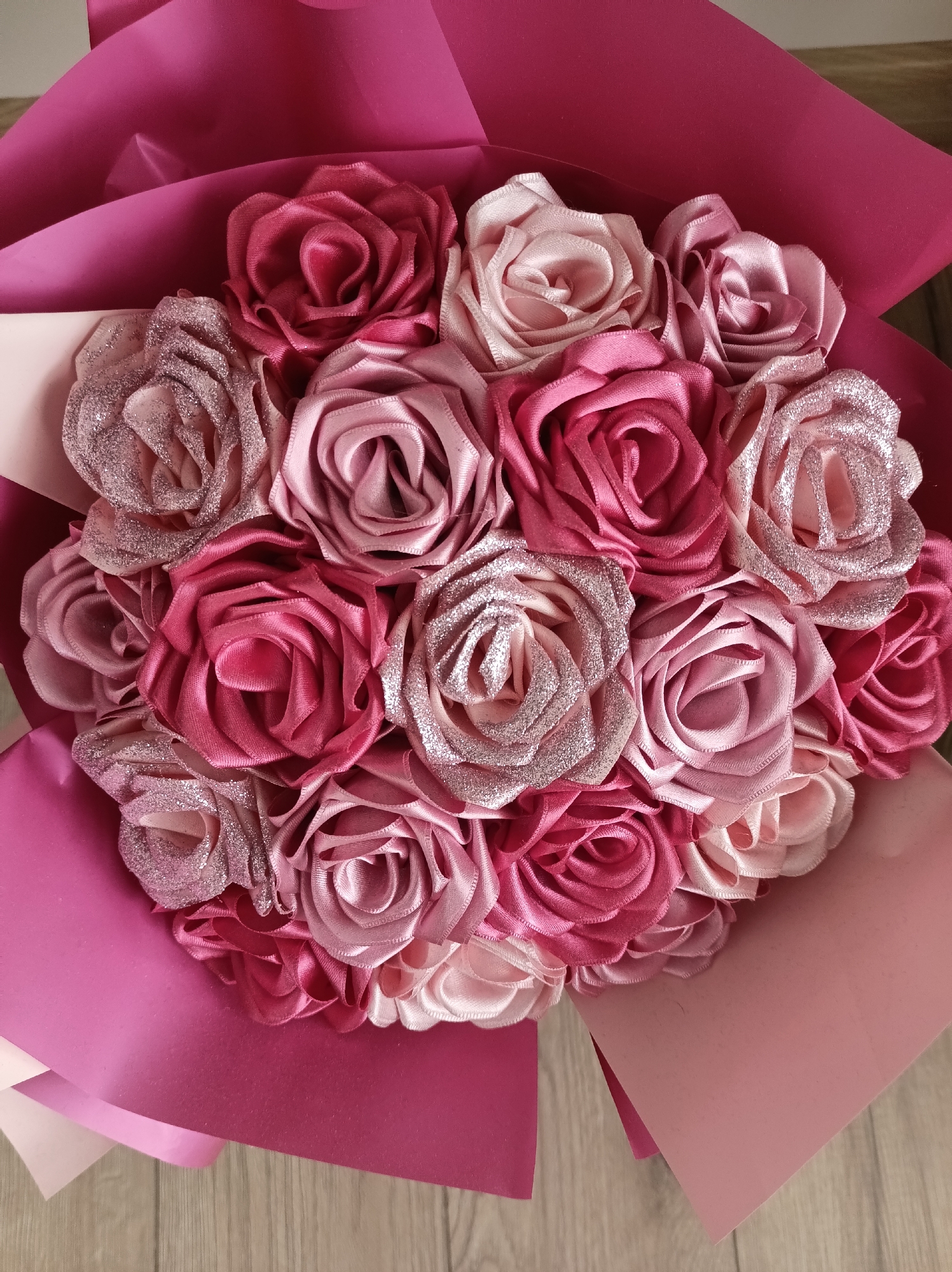 A bouquet of approximately 18 roses in varying shades of pink, light pink, and glittery pink is centrally positioned. The roses are arranged in a tight cluster, wrapped in several layers of pink paper. The arrangement is placed on a light-colored wooden surface. The scene contains no people or identifiable location details linking it to the city of Gabrovo, Bulgaria.