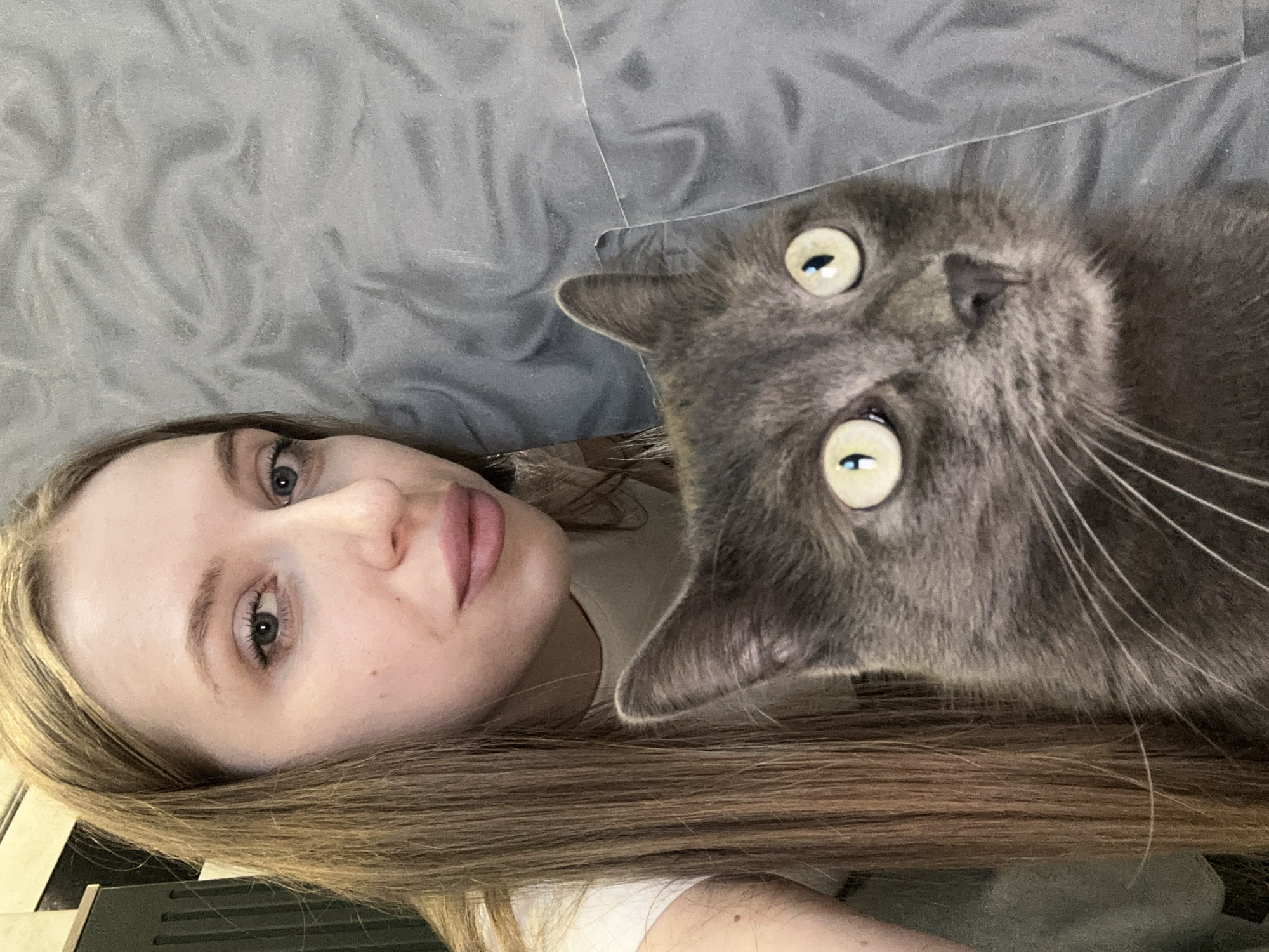 A person with light skin and long brown hair is lying on a gray textured surface, possibly a bed. Adjacent to them is a gray cat with large, round eyes and visible whiskers. The image is taken from a close-up, slightly overhead perspective.
I don't have location information.