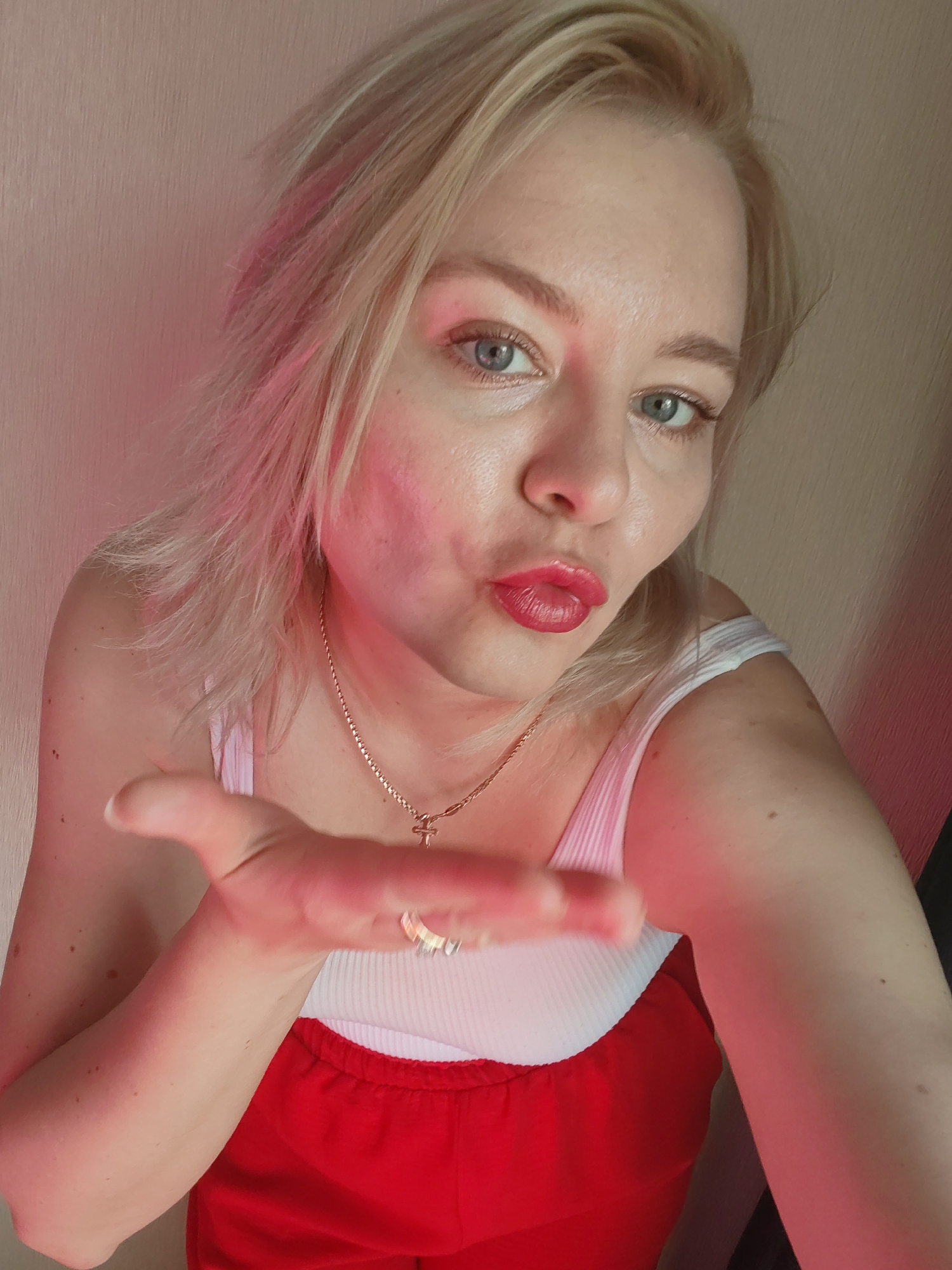 A blonde woman with light blue eyes is photographed close-up. Her face is partially illuminated by a pink light source. She is wearing red lipstick and making a kissing face. She is wearing a white tank top underneath a red dress. A gold necklace with a cross pendant is visible around her neck. She is extending her right hand towards the camera with an open palm, on which a ring is present. The background appears to be a light-colored wall. There is no information available to discern the location in Izhevsk, Russia.
