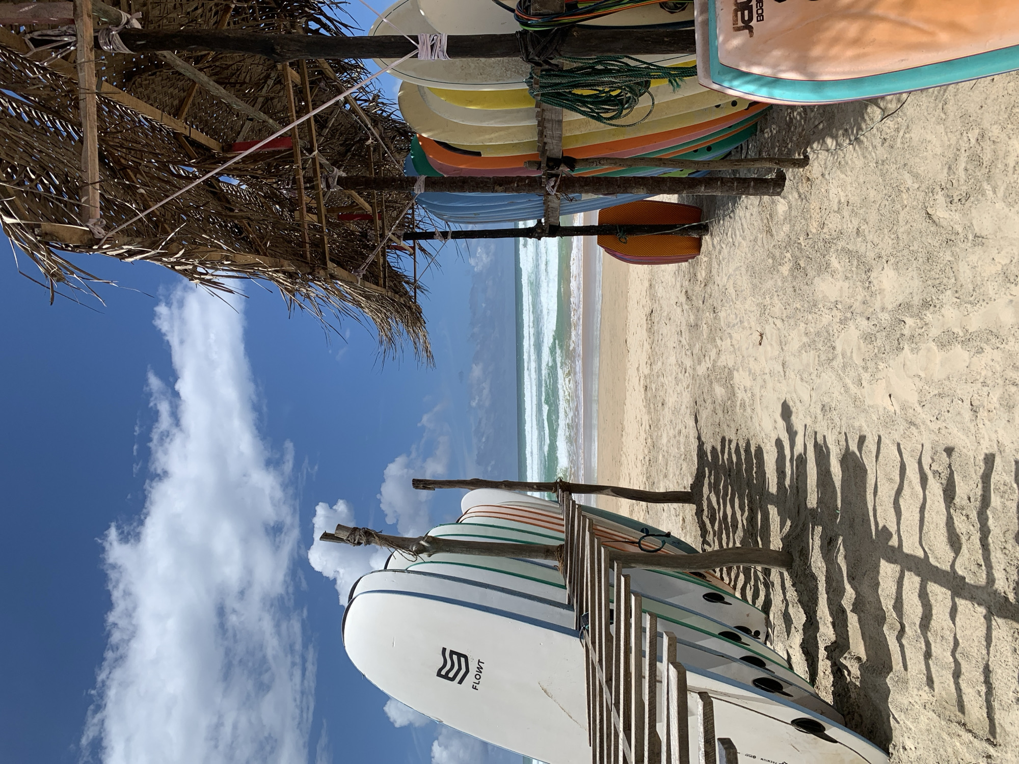 A row of surfboards stands on a sandy beach in Ahangama, Sri Lanka, under a thatched roof shelter. The surfboards are arranged in two groups. The first group, on the left, leans against wooden planks and is mostly white with colored stripes. The second group, on the right, is a vibrant array of colors, including blue, orange, yellow, and green. The ocean is visible in the background with gentle waves breaking on the shore. The sky is blue with scattered white clouds. The sand is light brown and shows shadows cast by the surfboards and the shelter. No people are visible in the frame.
