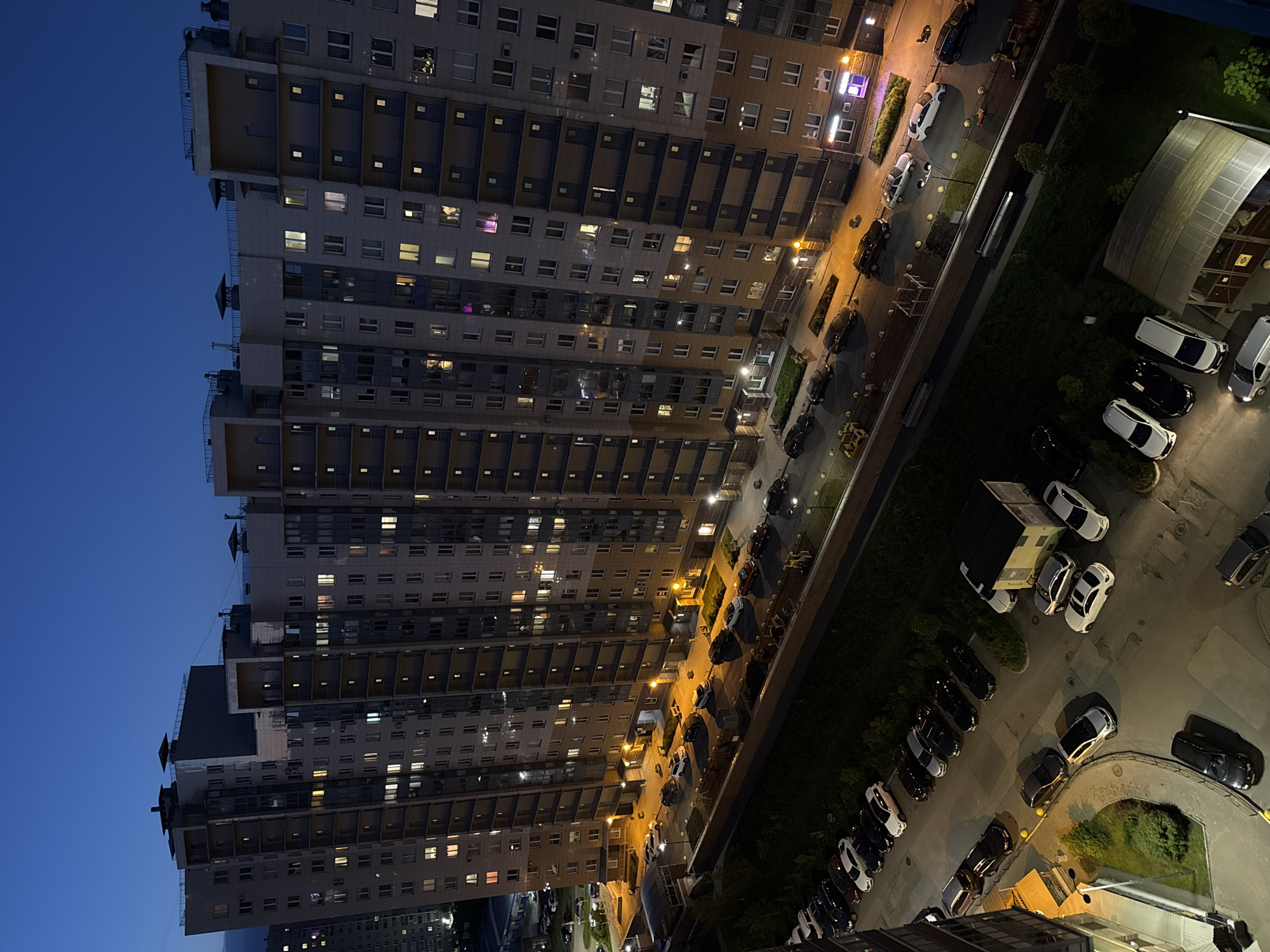 The image presents a high-angle, aerial perspective of a residential neighborhood at dusk or night. A tall, multi-story apartment building dominates the left and center of the frame, stretching diagonally across the composition. Many of its windows are illuminated, indicating occupancy within the numerous residential units.
Adjacent to the building is a street, well-lit by multiple streetlights that cast warm, yellow light onto the road and the surrounding green areas. A considerable number of cars are parked along the street and in various parking lots visible further to the right. These vehicles include sedans and SUVs, predominantly in shades of white, black, and silver.
In the mid-ground, a distinctive covered structure with a translucent, arched roof is visible, possibly serving as a carport or shelter. Near this, a dark, cube-shaped utility box or shed is situated amongst parked cars. Small patches of grass, bushes, and trees are interspersed between the parking areas and the street. Faint figures, possibly people or children, are vaguely discernible near what appears to be playground equipment in the upper-right portion of the scene.
The sky on the far left is a deep, clear blue, characteristic of twilight or early night. The scene is largely static, depicting a typical urban residential environment after sunset, with artificial lights providing the primary illumination. No discernible text is visible on any signs, buildings, or vehicles.