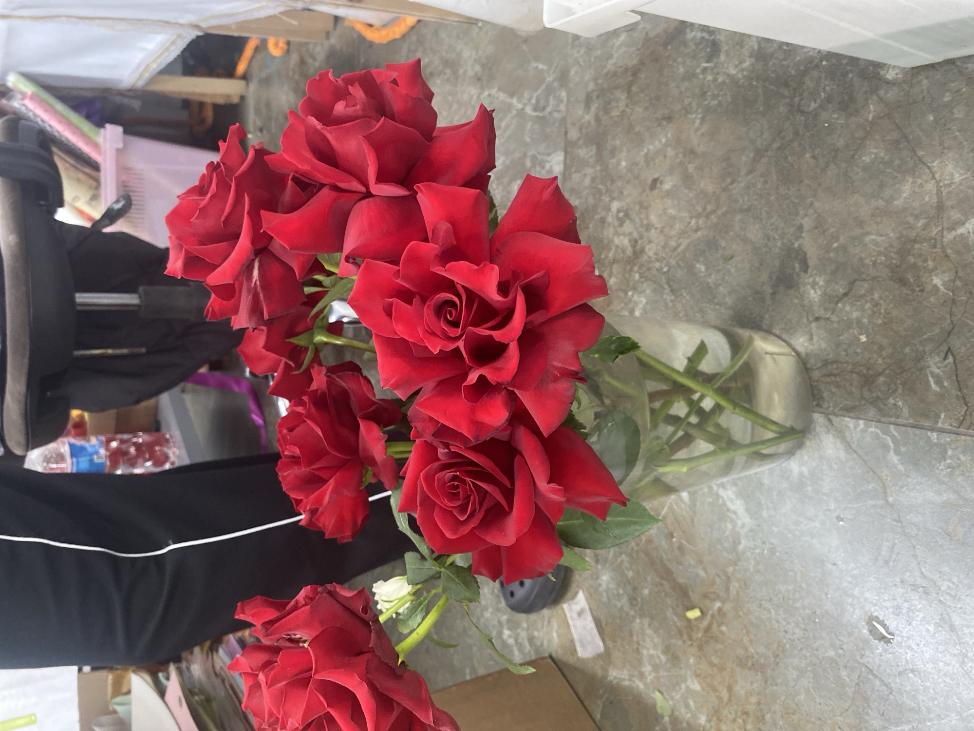 A vase of red roses sits on a grey, marble-patterned floor. The vase is glass and filled with water, stems, and green leaves. The roses are fully bloomed and appear to be freshly cut. In the background, there are various unidentified objects suggesting an indoor space, potentially a storage or preparation area. A black garment with a white stripe is visible on the left. A partial view of a baby stroller is also present. The specific location in Yekaterinburg, Russia is indeterminable based on visual data provided.