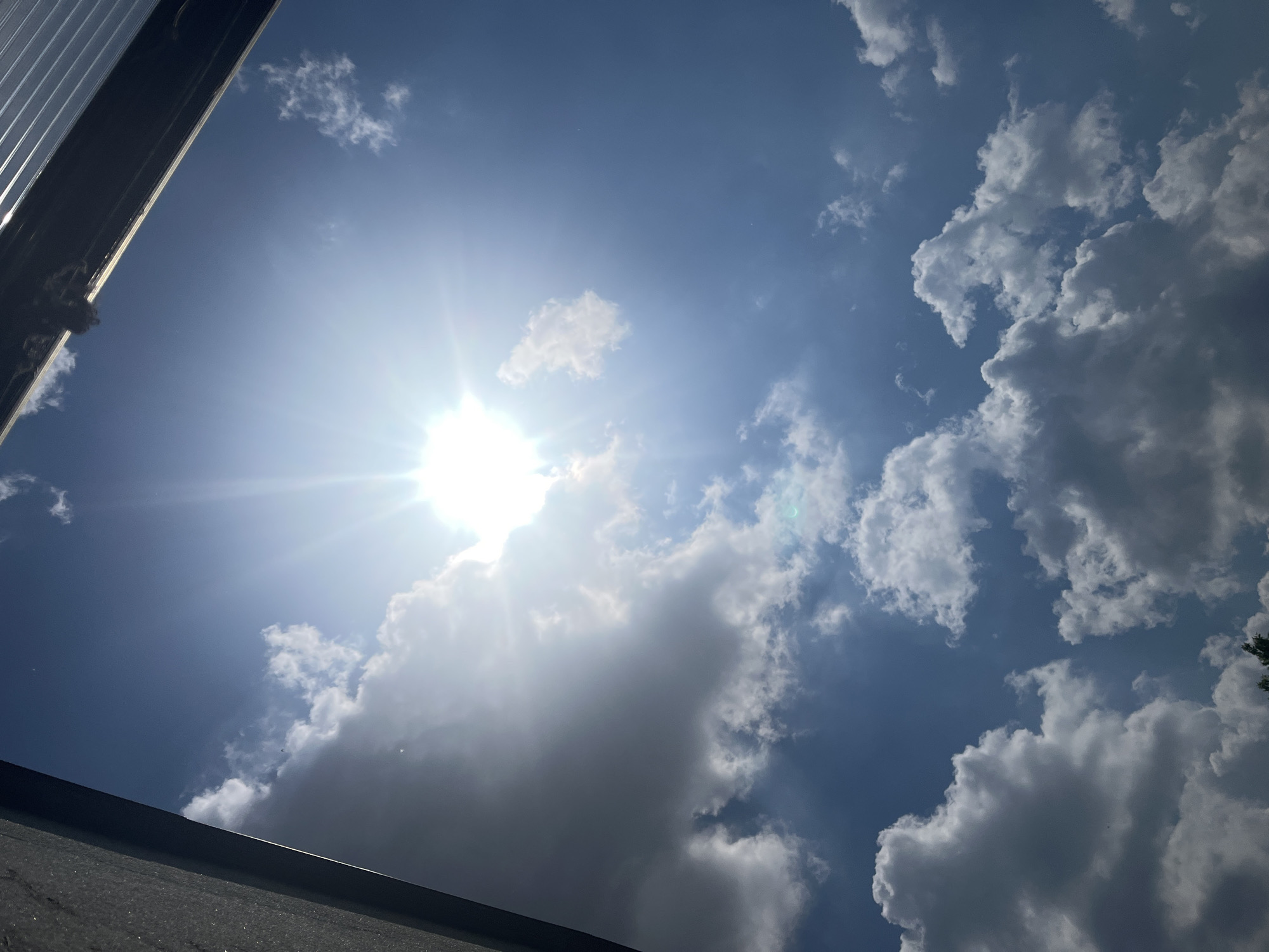 The image captures a bright, sunny daytime sky filled with clouds, viewed from a low angle looking upwards. The sun is a dominant feature, positioned in the upper left quadrant of the frame, emitting intense light and a prominent lens flare that spreads across the sky, particularly illuminating the clouds closest to it.
The sky is a vibrant blue, ranging from a lighter tone near the sun to a deeper blue in areas with fewer clouds. Various cumulus clouds are scattered across the sky. Some clouds appear brilliant white, brightly lit by the sun, especially those in the central and upper-right areas. Other clouds, particularly in the lower and right parts of the image, are darker and appear grey, indicating thicker cloud cover or being partially in shadow.
In the bottom-left and top-left edges of the main image, and more clearly in some crops, a dark, angular structure is visible. This appears to be part of a building or roofline, possibly made of metal or asphalt, suggesting the photo was taken while looking up from a position near a structure. No people or specific objects are identifiable within the scene, apart from the natural elements and the architectural edge. There is no visible text. The overall impression is one of a clear, bright day with a mix of sun and clouds.