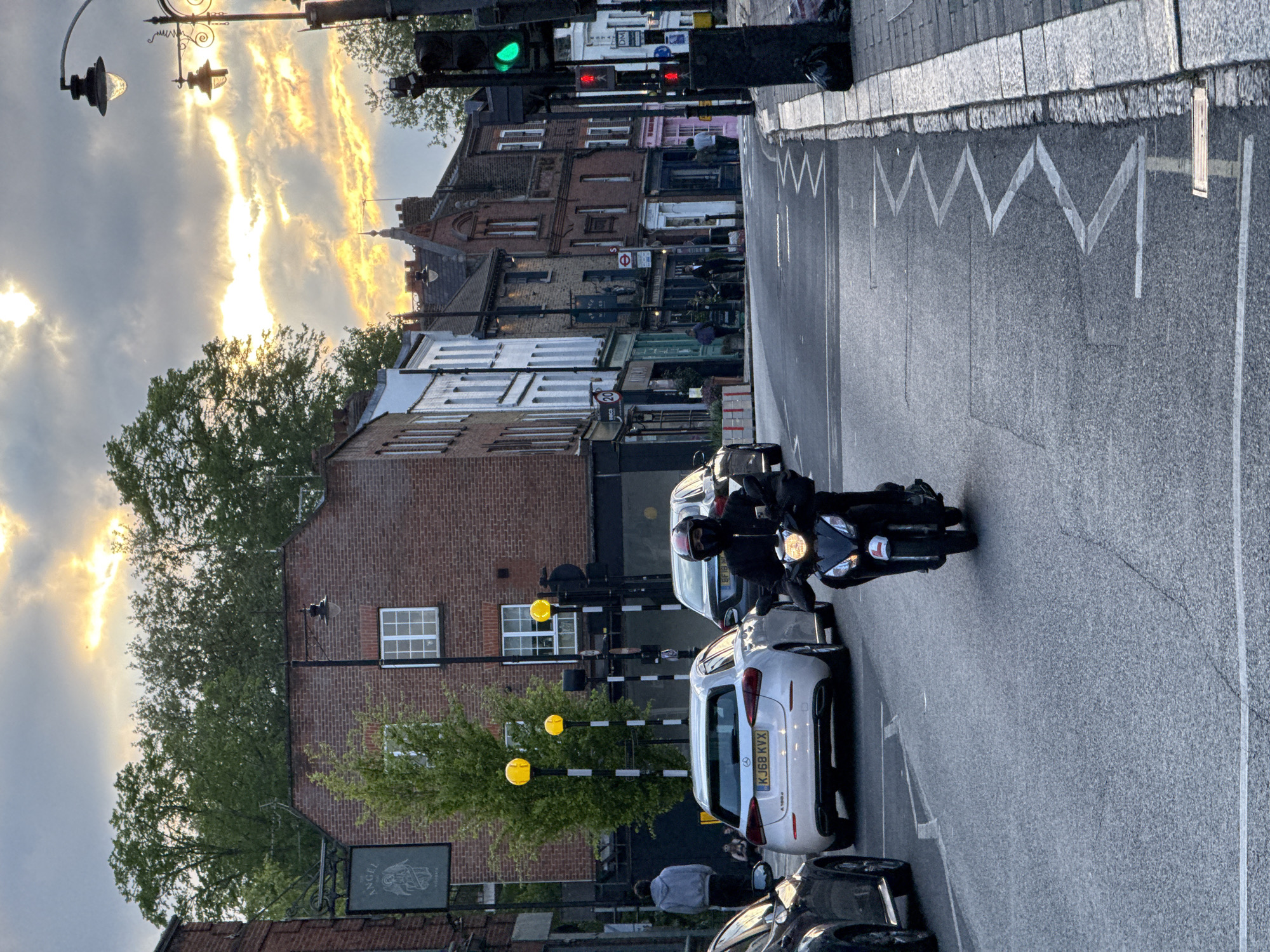 The image shows a street scene in London, United Kingdom. The view is taken from a slightly elevated position, looking down onto a road intersection. The road runs away from the viewer with a slight curve to the right.
A motorcycle with a rider is positioned near the center of the frame, close to a white Mercedes car. Another car, darker in color, is parked behind the white car. The traffic lights closest to the viewer are green, while lights further up the road are red.
The buildings lining the street are a mix of brick and painted facades. A pub sign, "The Angel", is visible on the left side of the frame. Sidewalk markings, zig-zag lines, are noticeable on the right side of the road.
In the sky above the buildings, the light shows sunset, giving the scene an overall warm-toned feeling.