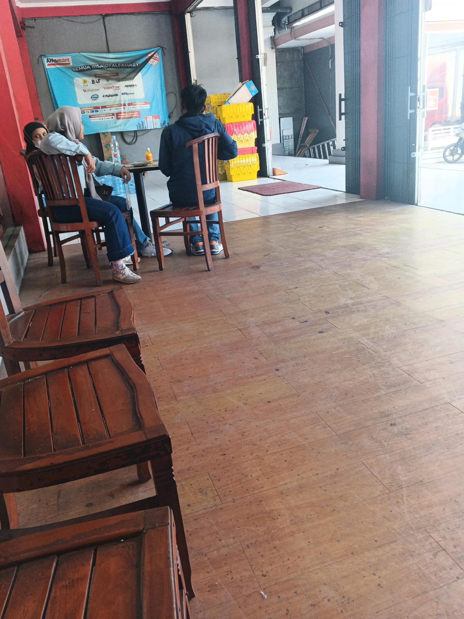 In Genuk Barat, Indonesia, three individuals are seated at tables in an establishment that appears to be a convenience store or cafe. One person, wearing a hijab and a light blue top, is seated at a table. Another person, wearing a dark blue hoodie, is seated at the same table. A third individual, partially visible and wearing a black head covering, is seated at a separate table. Wooden chairs surround the tables, and the floor is made of polished wood planks. A blue banner advertising "ALFAMART" is visible on the wall, indicating the location is likely an Alfamart store. The overall scene depicts a casual waiting or resting area within a commercial establishment.