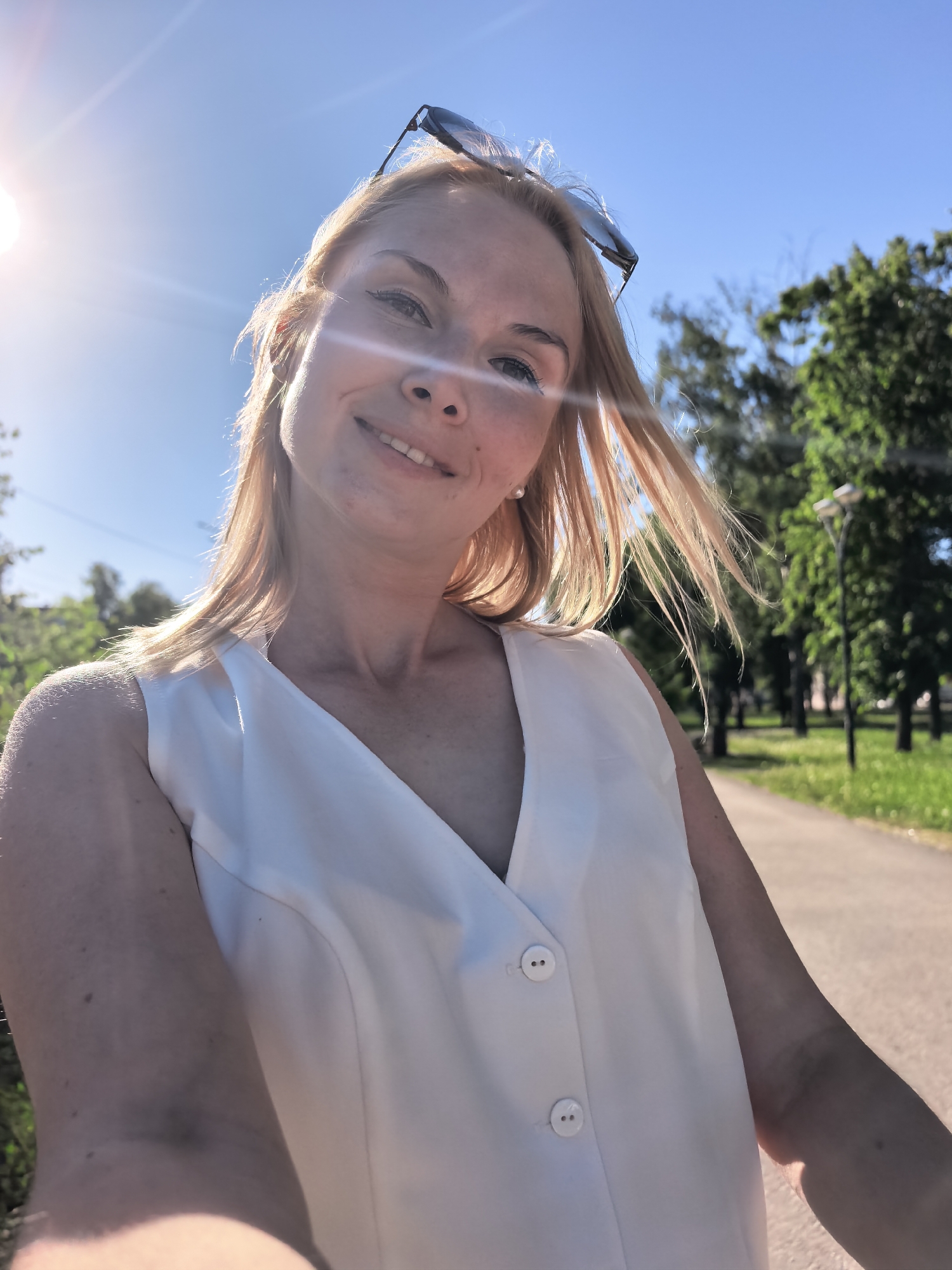 A selfie of a blonde woman in a white sleeveless button-down shirt, wearing sunglasses on top of her head. She is smiling. The location appears to be a park in Sterlitamak, Russia, indicated by the presence of a paved path, trees, and streetlights in the background. The sky is blue, and there is bright sunlight.