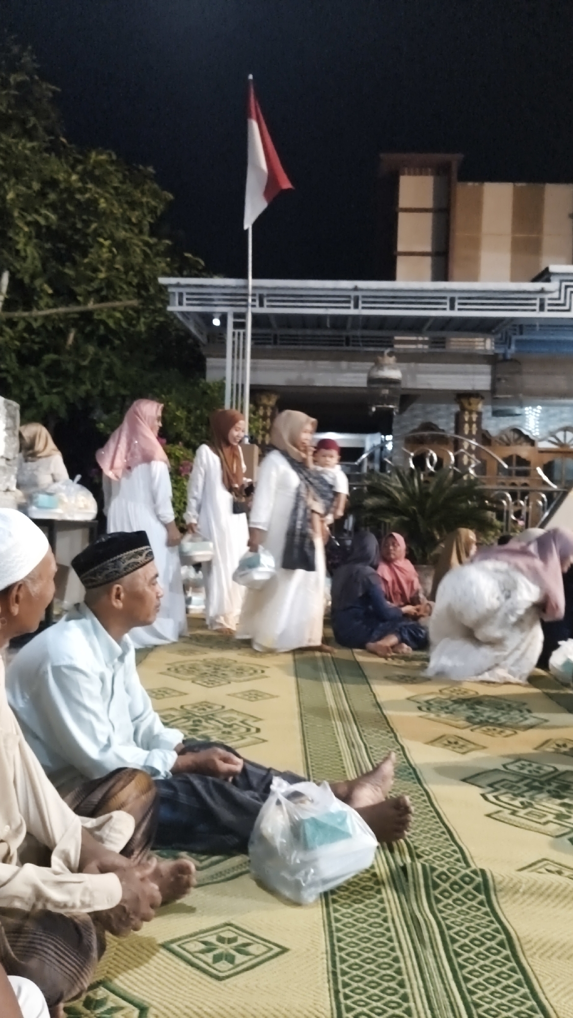 In the city of Warulor, Indonesia, a gathering is taking place outdoors at dusk or night. The scene is dominated by people dressed in traditional attire, with many women wearing hijabs. In the foreground, several men are seated on a mat with a green and yellow geometric pattern. One man on the left is partially visible, wearing a light-colored garment and a white skullcap. Next to him, a man wearing a light blue long-sleeved shirt and a black peci (cap) is seated, with a plastic bag containing items beside him. Further back, several women are standing and interacting. One woman in a pink hijab is holding a white bag. Another woman in a brown hijab is smiling, and a third woman in a cream-colored hijab is holding a baby. Other individuals, including children, are seated on the mat in the middle and background of the scene. The environment includes lush green trees on the left and a building with a distinctive facade featuring horizontal and vertical banding in the background. A flag, which appears to be the Indonesian flag with red and white colors, is prominently displayed on a pole, adding a patriotic element to the gathering. The lighting suggests an evening event, possibly a celebration or religious ceremony.