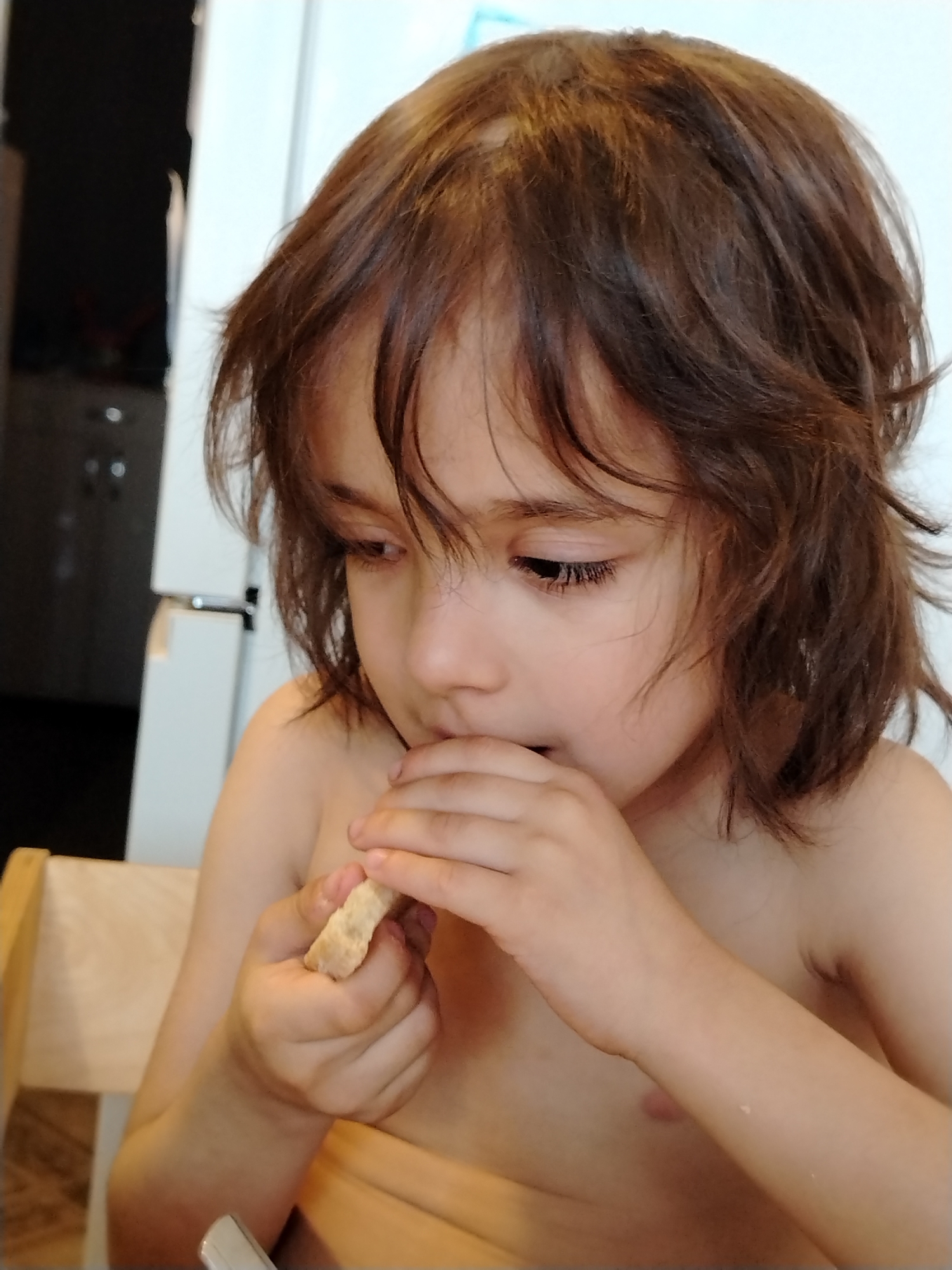 This is a close-up, vertical, eye-level shot of a young child, possibly a girl, with medium-length, brown, slightly wavy hair. The child is shirtless and sitting at a wooden table, as indicated by the edge of a wooden chair on the left side of the frame. The child is looking down and to the right, with their hands raised to their mouth, and appears to be eating a small, light-colored piece of food, possibly a cracker or biscuit. The child's expression suggests focus or concentration. The background is out of focus, showing a white wall on the right and a dark area with vertical lines on the left, which could be a kitchen appliance or furniture. The lighting suggests it is daytime, likely indoors. There is no visible text in the image.