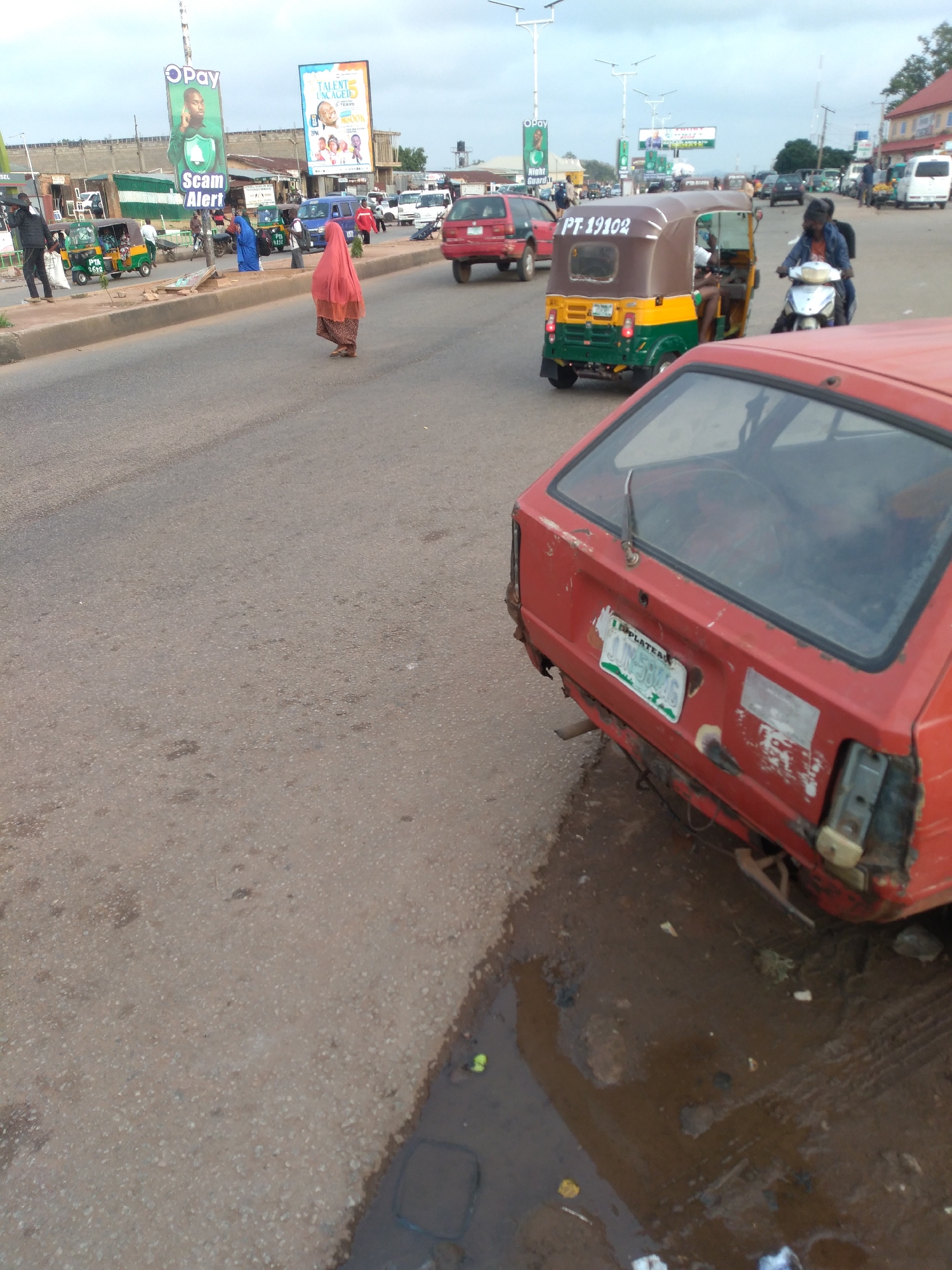 In Gura Top, Nigeria, a street scene unfolds with a reddish-brown asphalt road and a concrete median. A woman in a pink veil walks across the road, while a green and yellow rickshaw, marked "PT 19102," navigates traffic. A motorcyclist in a blue jacket rides past. Further down the road, a red car with a damaged bumper and a Nigerian license plate, "GN-58MG," is parked. Billboards stand along the roadside, one advertising "OPay" and another with "Talent Uncaged." The sky is overcast, and the environment suggests a bustling urban area with a mix of vehicles and pedestrians.