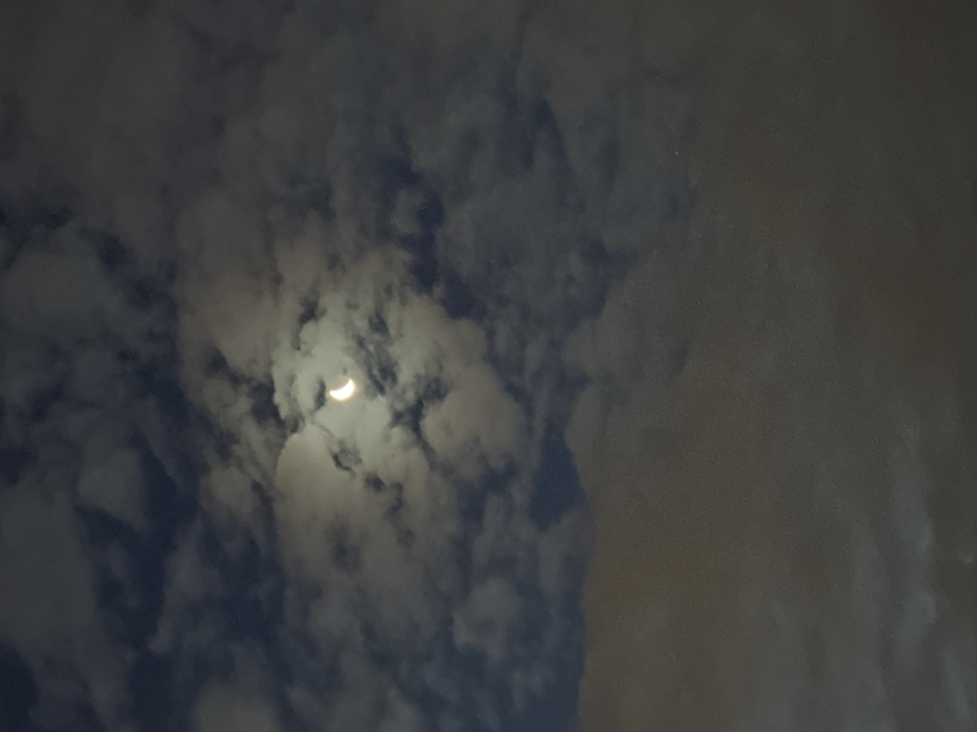 The image depicts a crescent moon partially obscured by clouds at night. The sky is filled with various cloud formations, some dark and dense, others wispy and illuminated by the moon's light. The moon itself is a thin sliver, bright and distinct against the darker parts of the sky. The lower portion of the image is dominated by a large, blurry band of clouds, appearing more opaque and darker than the clouds surrounding the moon. The overall impression is of a moody, atmospheric night sky. There are no discernible landmarks or features that would indicate a specific location within Tallinn, Estonia, or any other city. The image solely focuses on the celestial event.