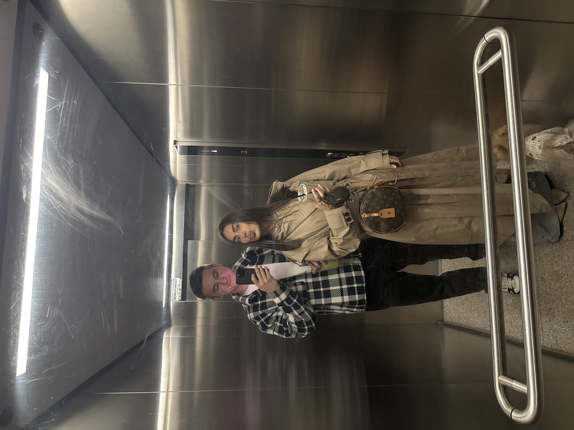 Two individuals are photographed in an elevator, presumably in Warsaw, Poland, as indicated by a sign in Polish visible in the mirror. One individual is male, wearing a black and white checkered shirt and black pants, holding a phone taking the picture. The other individual is female, wearing a tan trench coat and black pants. She is carrying a brown purse. The elevator is metal and features a handrail.