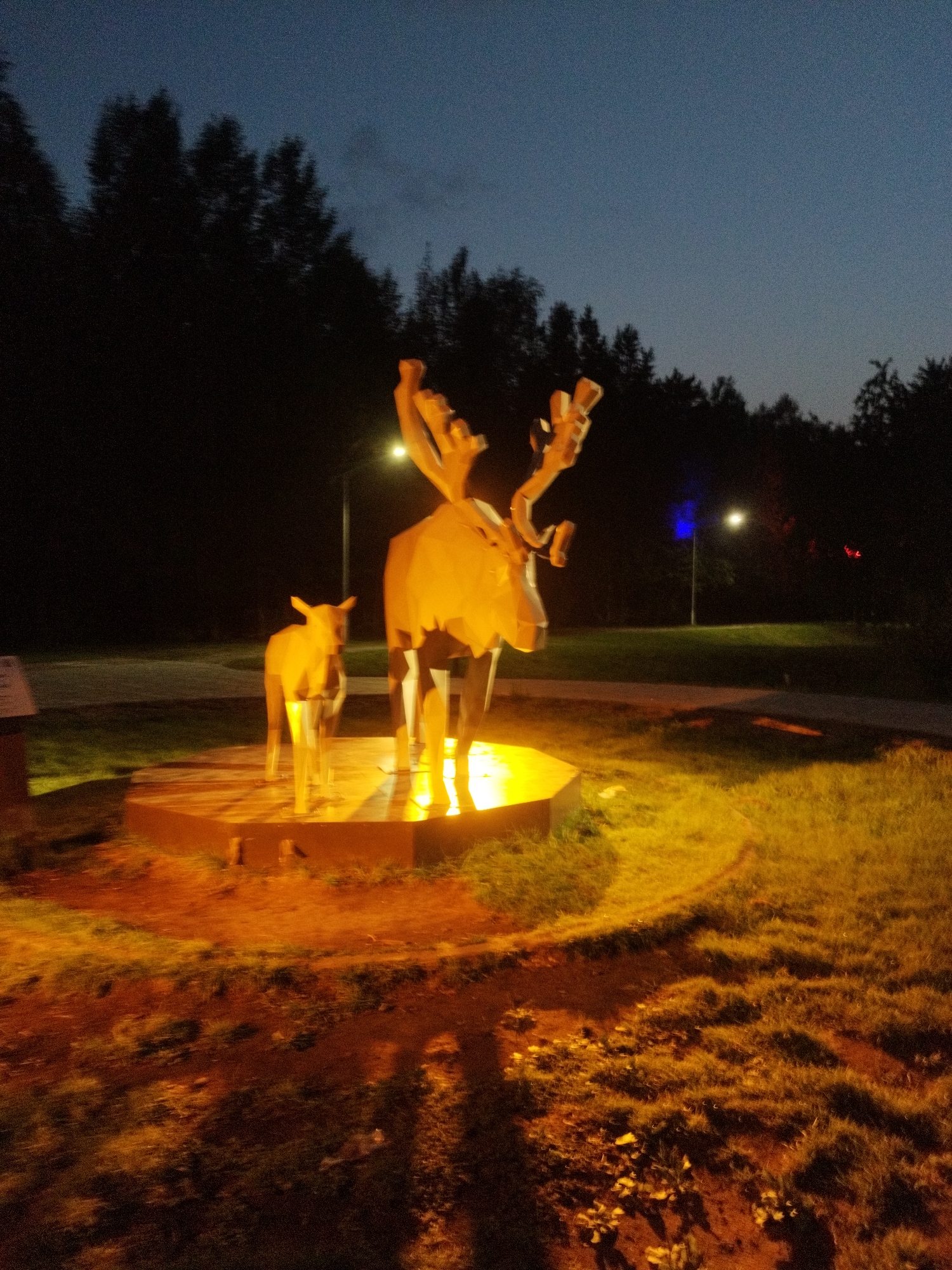 The image depicts a twilight scene with two metal sculptures of reindeer, one adult and one calf, illuminated by yellow lights from below. The sculptures are positioned on a circular, raised platform. The background features a dark forest line against a twilight sky. In the mid-ground, there is a paved path and a lamppost emitting a blue light, suggesting an urban or park setting. The foreground consists of grass and soil, also illuminated by the warm light from the sculptures. The overall impression is that of an outdoor art installation in a park or public space. The location is identified as the Park of Culture and Rest "Surgut", Russia.