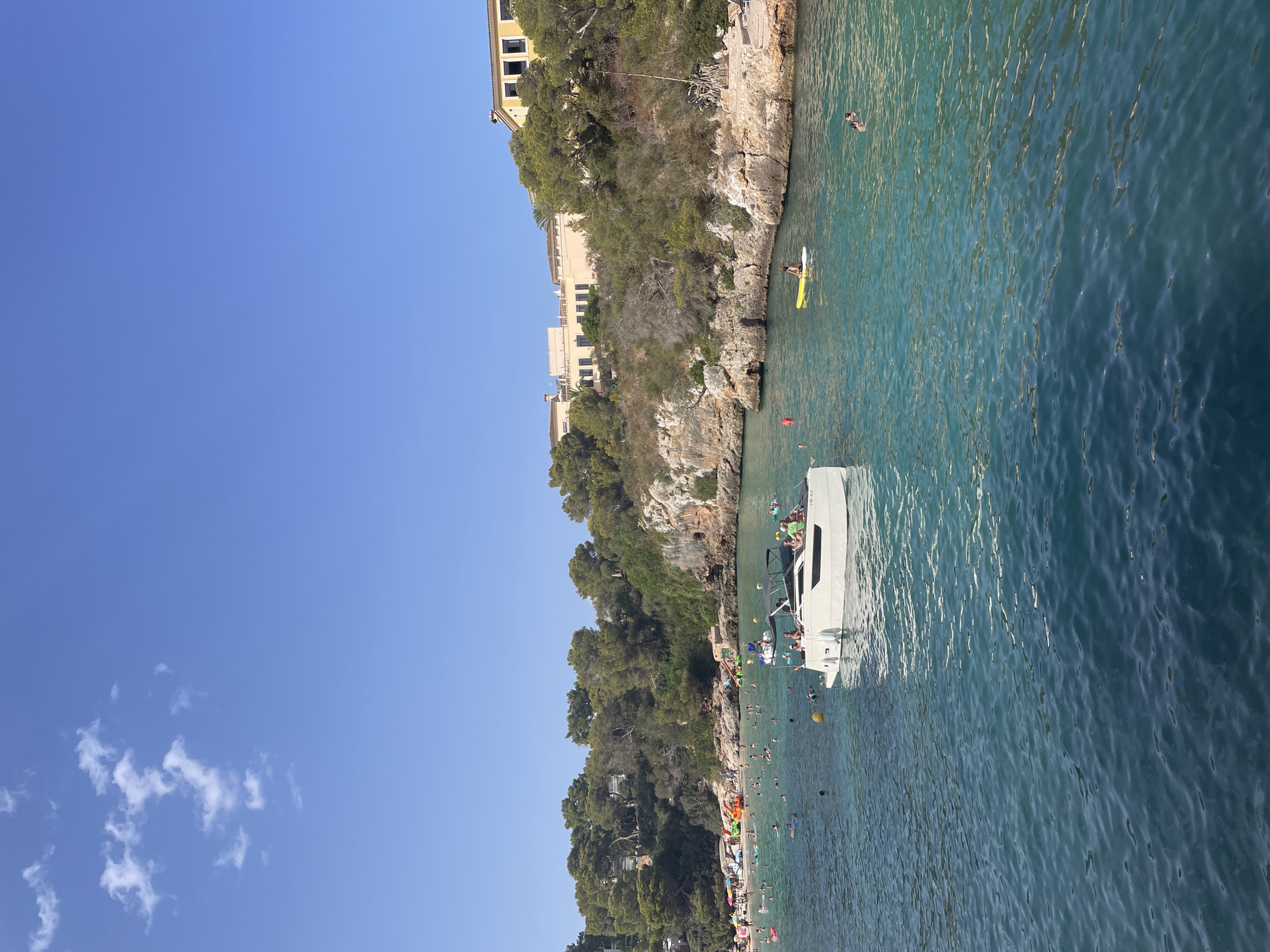 A wide-angle shot captures a bright, sunny day at Cala Ferrera, Spain. The clear blue sky is partially obscured by a few wispy white clouds. The foreground is dominated by the tranquil, turquoise waters of the Mediterranean Sea, with gentle ripples on the surface.

In the middle ground, a white motorboat with a black canopy cruises on the water, carrying several people. Nearby, individuals are engaged in various water activities. Some are swimming and floating on inflatable rings, while others are seen from a distance on the crowded beach. A yellow paddleboard with a person on it is visible further out in the water.

The background reveals a picturesque coastline characterized by rocky cliffs covered in lush green trees and shrubs. Several buildings with yellow and cream-colored facades are nestled on the hilltops, overlooking the bay. The beach itself is teeming with people enjoying the sun and sea. The overall scene depicts a vibrant and lively summer day at a popular coastal resort.