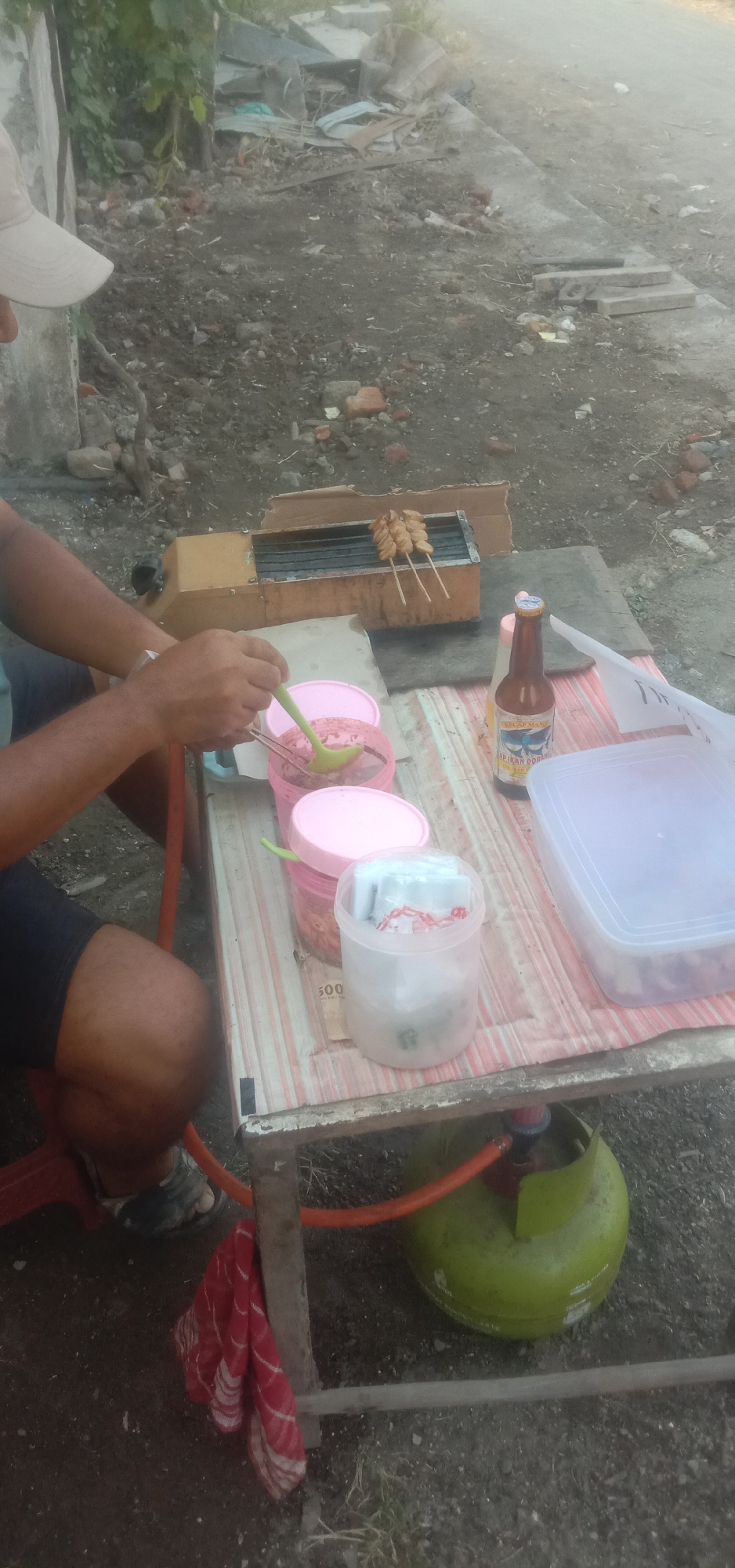 The image depicts an outdoor scene in Banjar Dowo, Indonesia. A person is seated at a portable table, preparing food. On the table, there is a small grill with several skewers of food cooking, two pink plastic containers with lids, one open and holding a red sauce with a green ladle, and another closed. A bottle of what appears to be soy sauce is also on the table. To the right of the containers is a clear plastic storage box. Underneath the table, a green gas cylinder with an orange hose is visible, along with a red and white patterned cloth hanging from the table leg. The ground is covered in dirt and debris, with some vegetation in the background. The overall impression is of a casual, makeshift food stall operating outdoors.