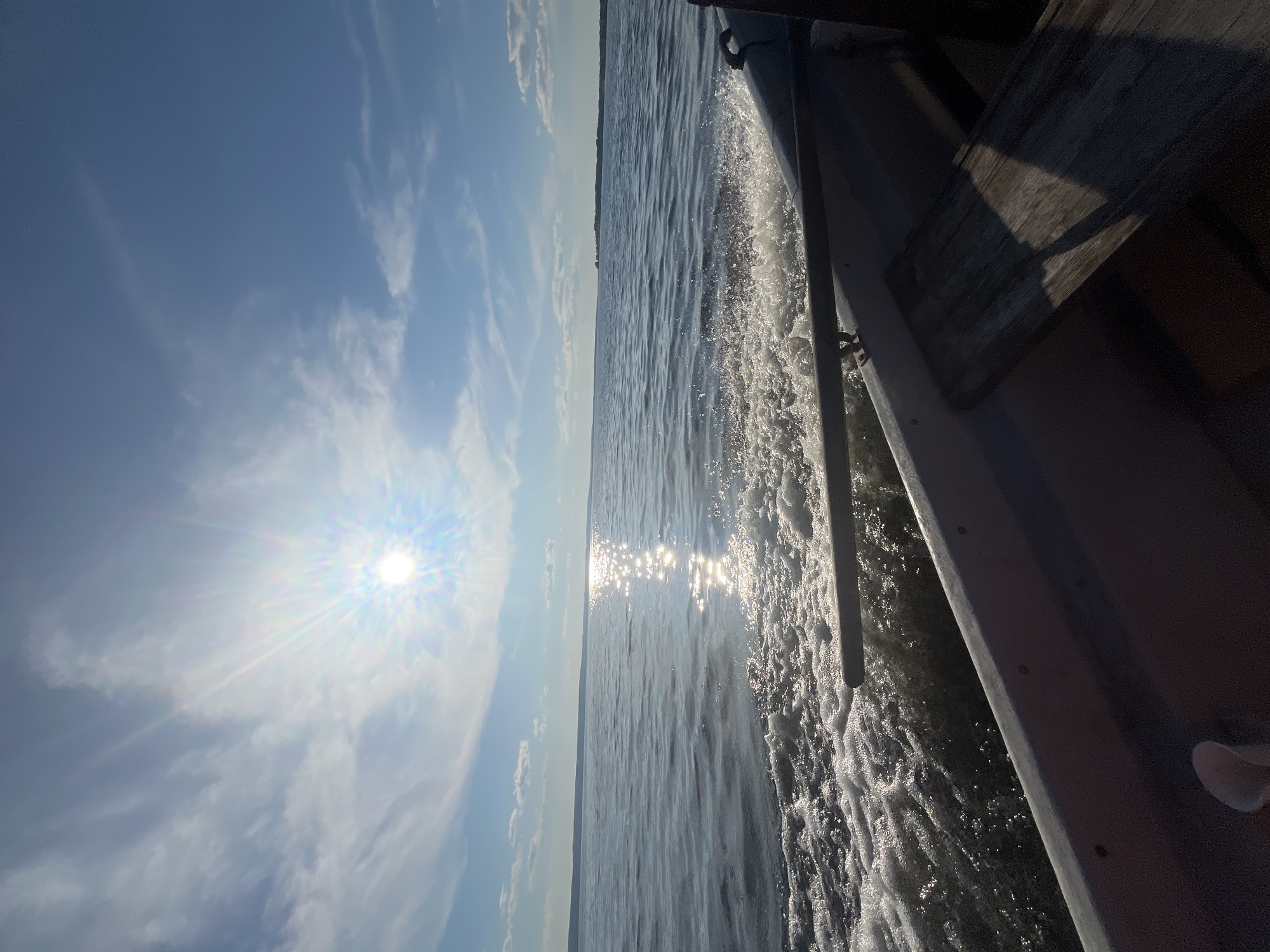 The image captures a view from inside a small boat moving across a large body of water, likely a lake or a calm sea. The sun is high in the sky, bright and positioned to the left, casting a strong glare on the water's surface and creating a shimmering reflection. Scattered white clouds dot the bright blue sky. The water is rippled, with a visible wake trailing behind the boat, indicating forward motion. A dark-colored oar or tiller extends across the foreground, partially submerged in the wake. The interior of the boat is visible in the lower right portion of the image, revealing a dark hull and a wooden plank. In the distance, a faint, tree-lined horizon is visible. The scene suggests a sunny day, possibly in the summer, experienced from a boat on a body of water near Siukterka, Russia.